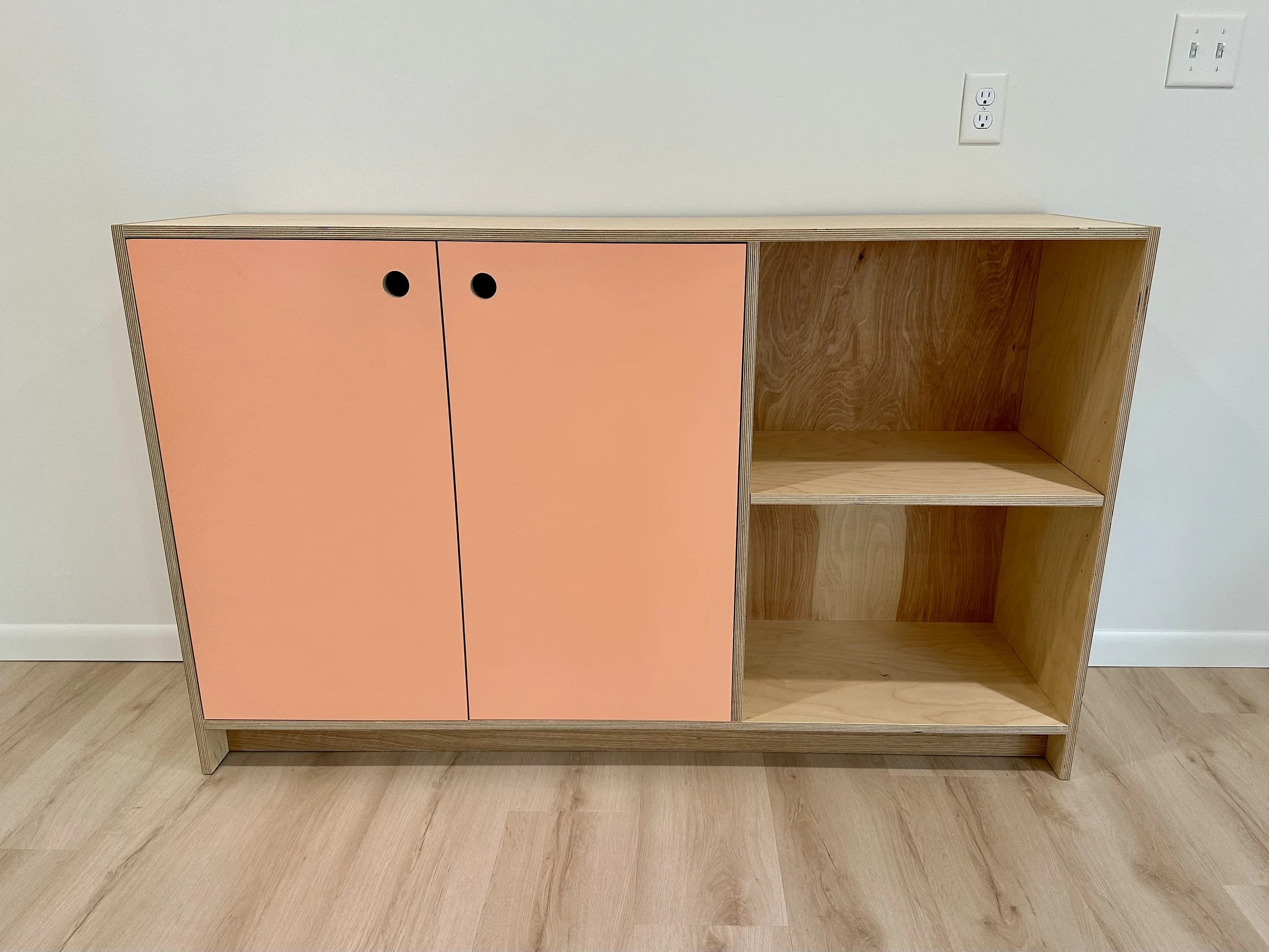 Wooden cabinet with two peach-colored doors on the left and open shelves on the right, set against a white wall with electrical outlets.
