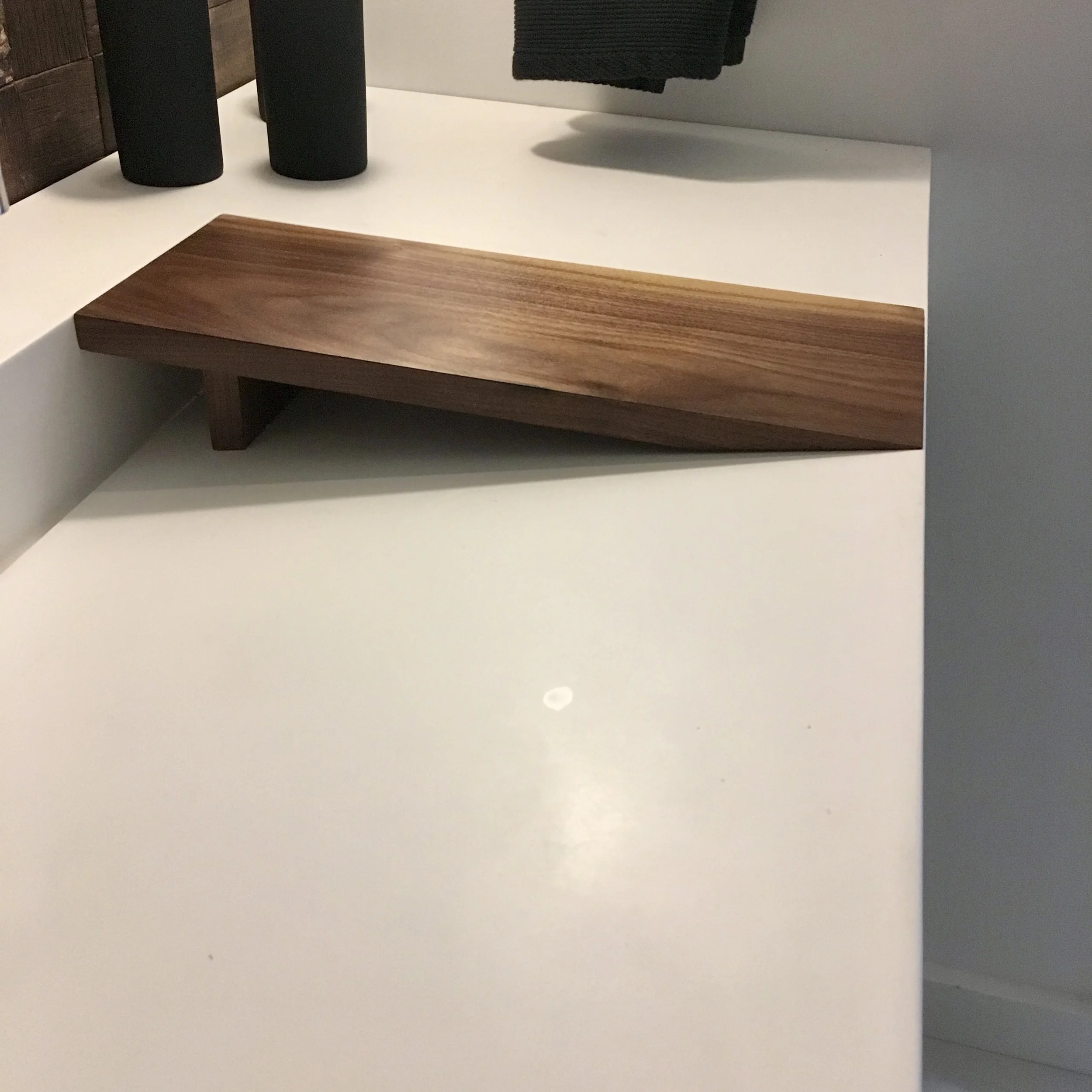 A minimalist wooden tray resting on a white surface with a black towel hanging above and two black cylindrical objects nearby.