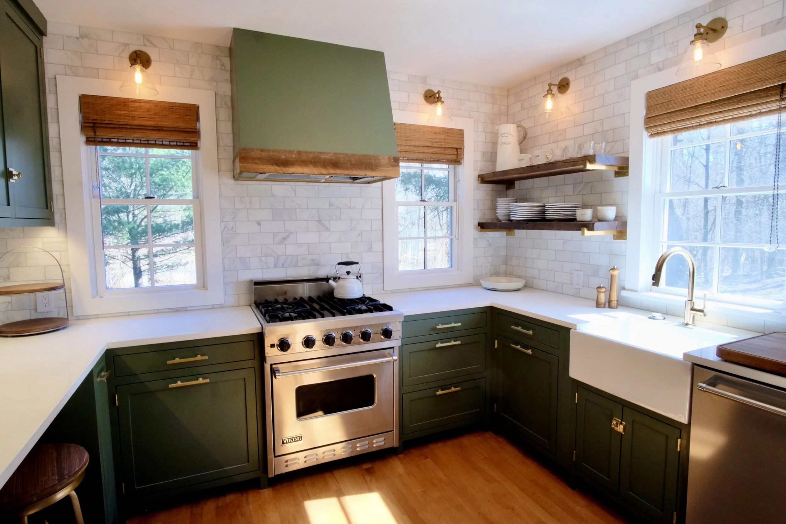Kitchen with green cabinets, white brick walls, wooden open shelves, stainless steel oven, white farmhouse sink, open windows with blinds, and warm lighting.