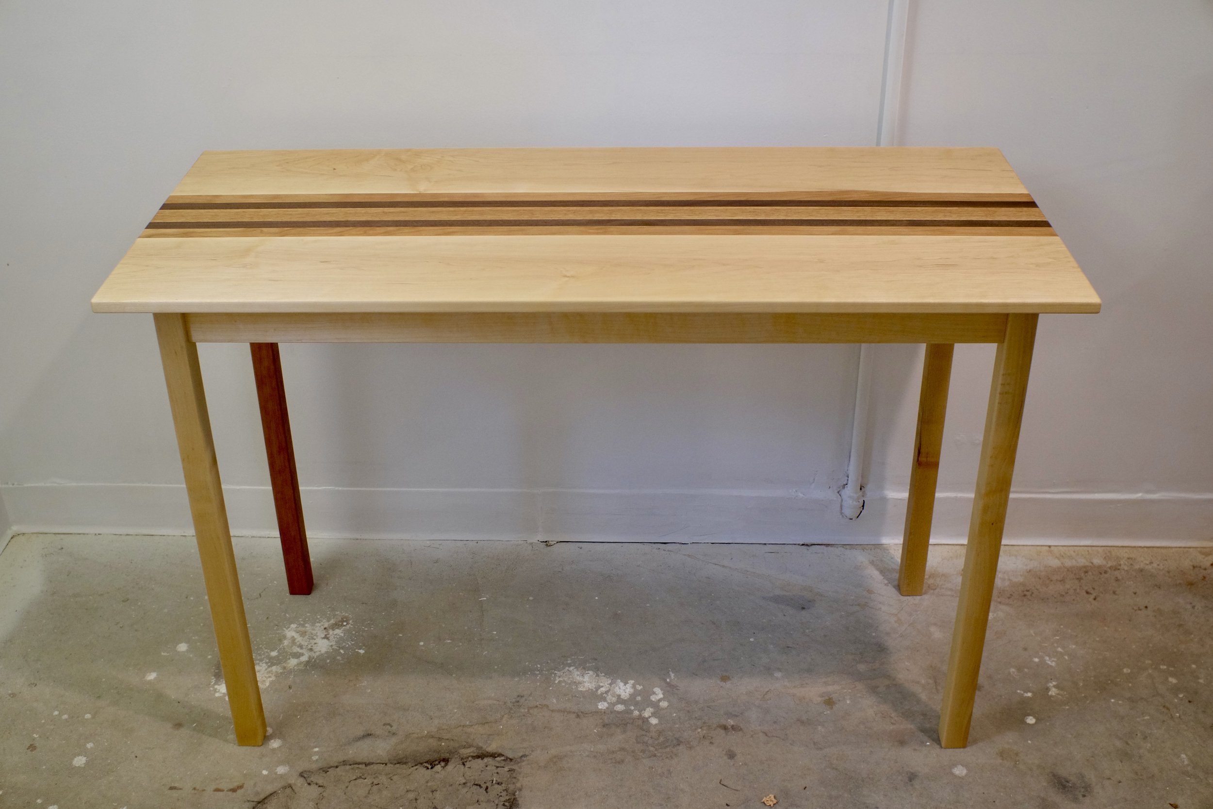 A wooden table with four legs, two of which are painted yellow, and a top with two dark stripes running across it, placed against a white wall with visible piping on the right. The floor appears to be concrete.