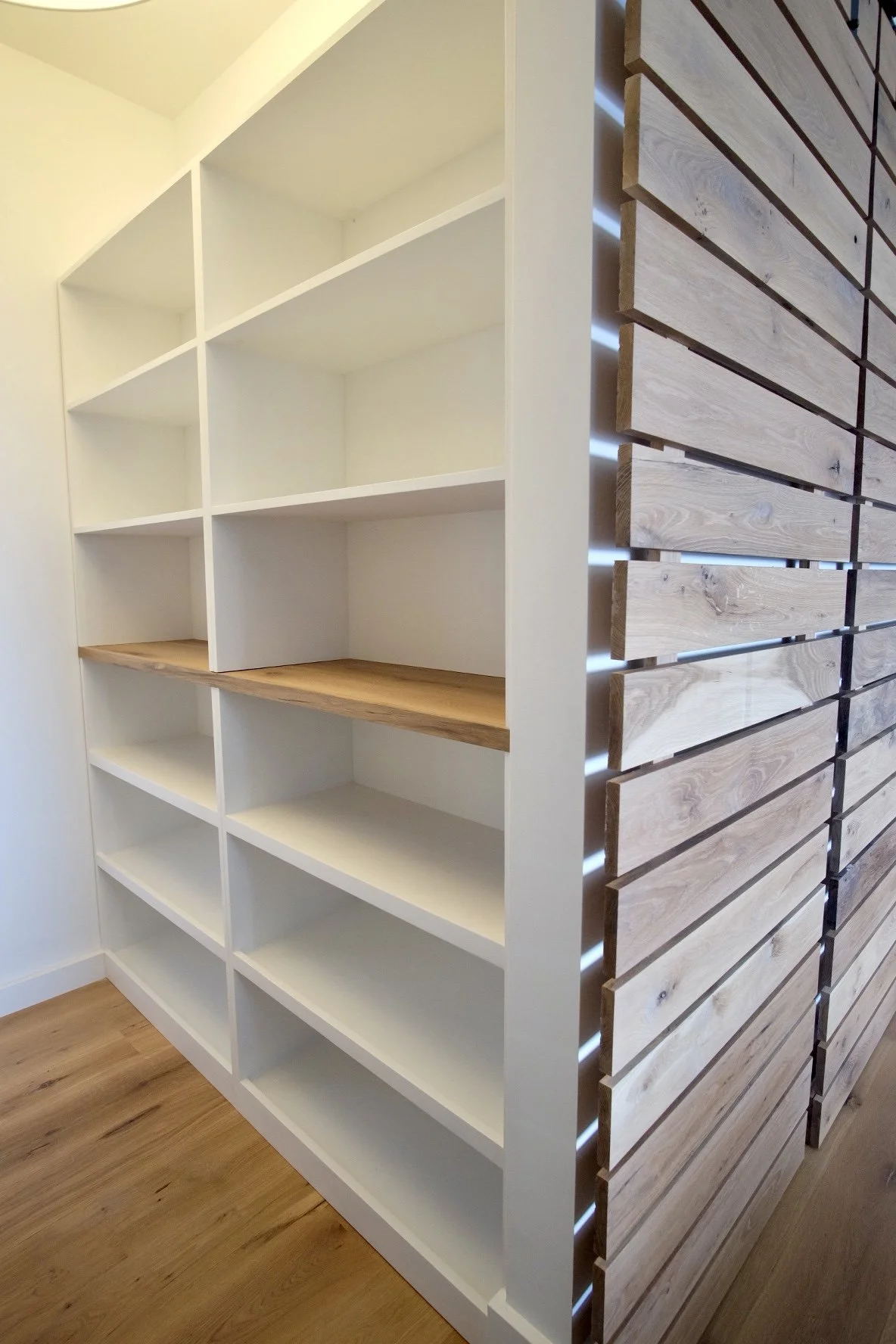 Empty built-in shelving unit with white shelves and a wooden countertop on the left side, and a wooden slatted wall on the right side, in a room with wooden flooring.