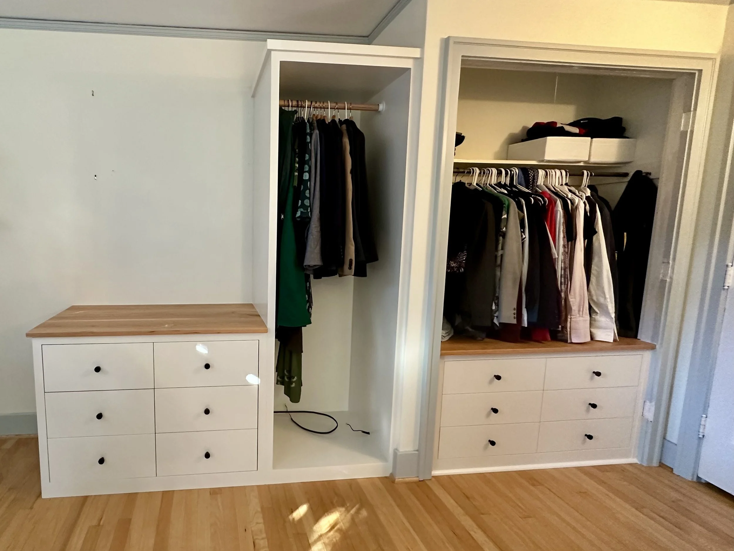 Open custom closet with clothes hanging on racks, shelves with folded clothes, and white drawers with black knobs, against a light-colored wall and hardwood floor.