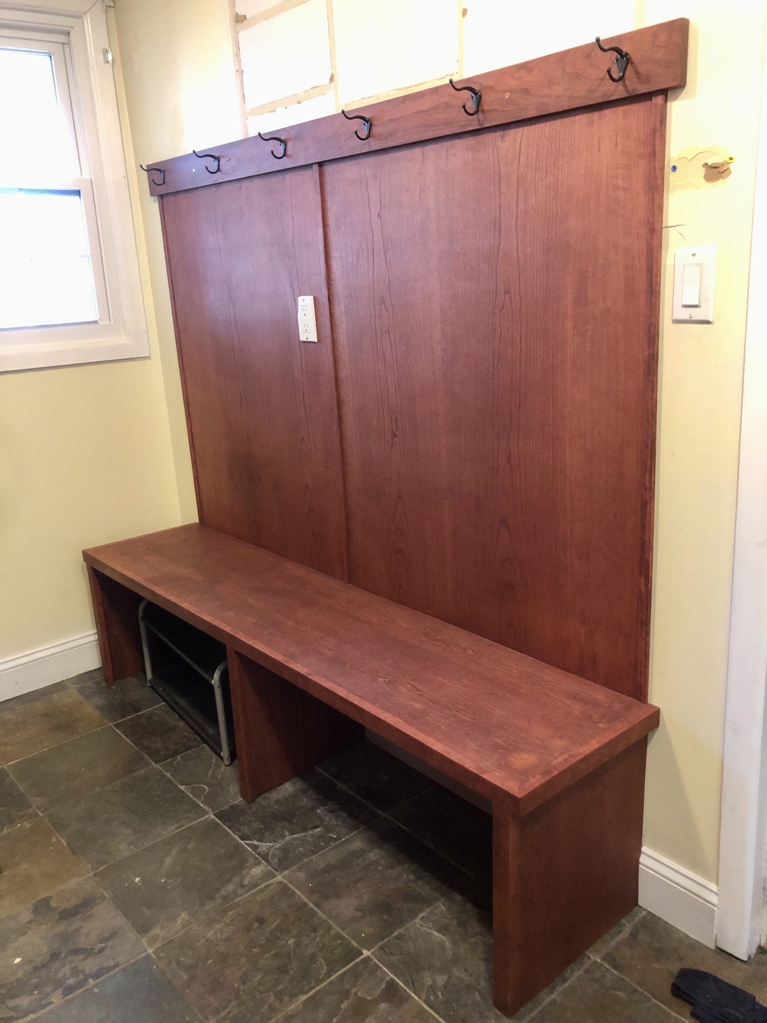A wooden hall tree with hooks and a bench, positioned next to a window on a tiled floor.