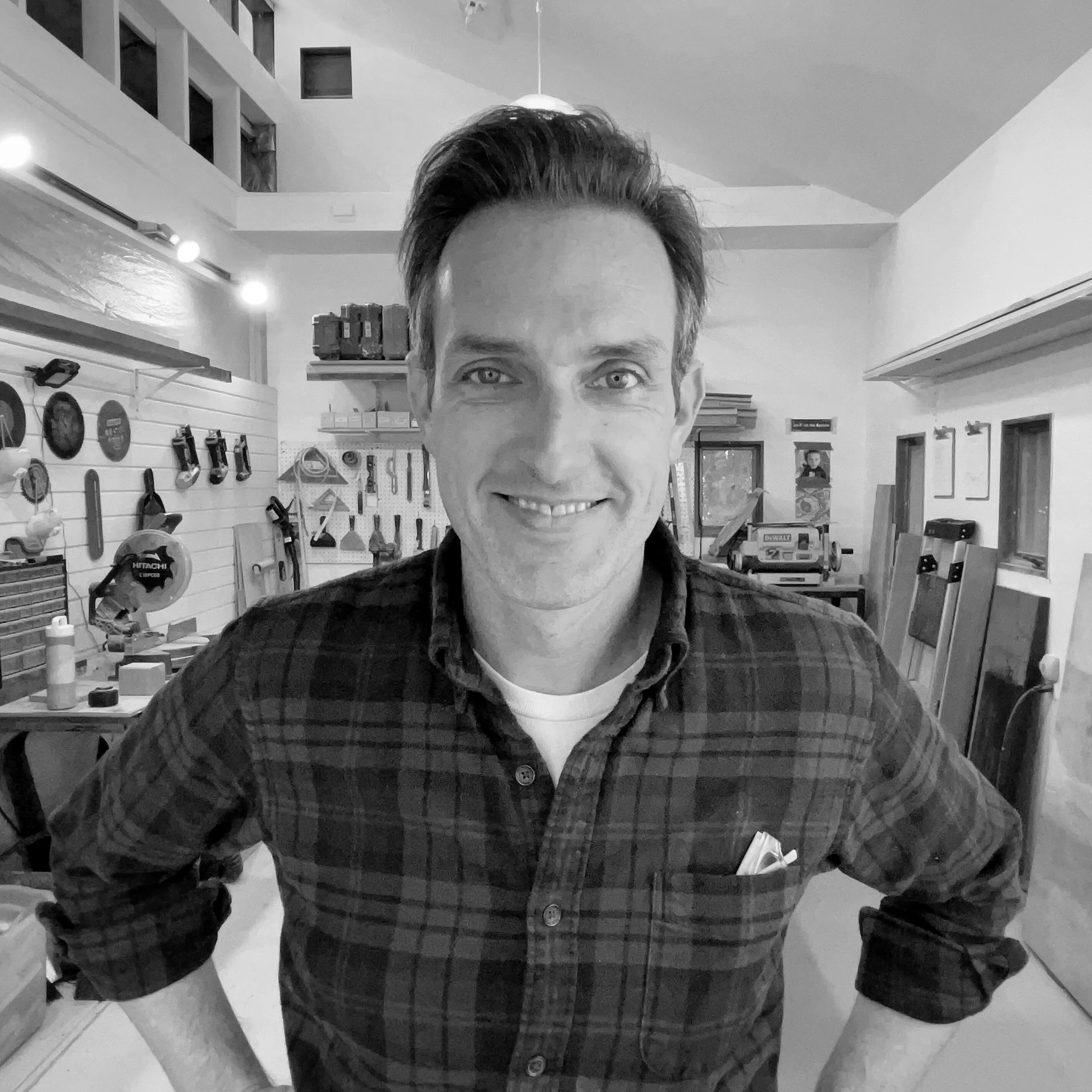 A smiling man with short hair, wearing a plaid shirt, standing in a woodworking workshop with tools and wood stored on the walls and shelves.