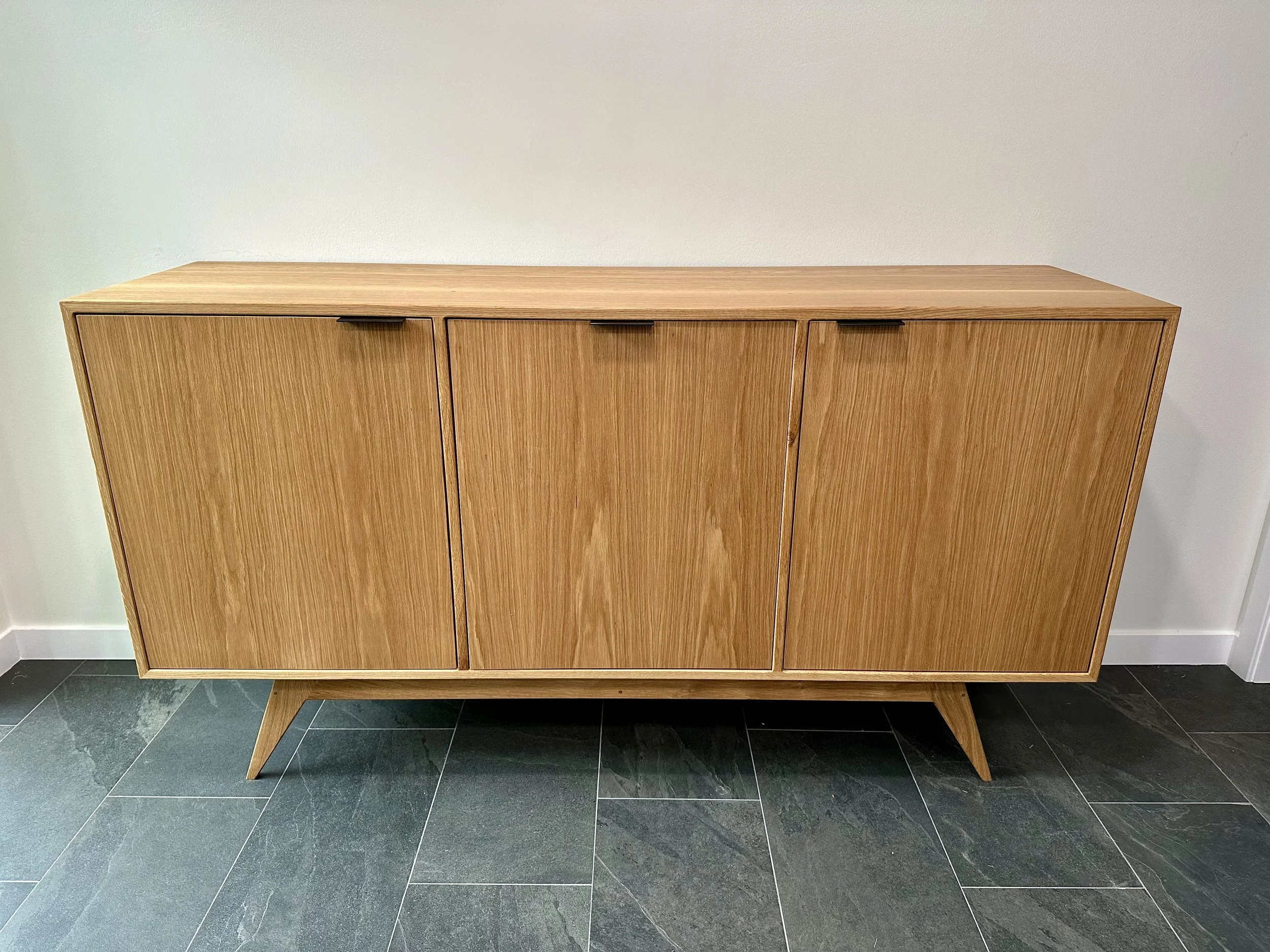 A wooden mid-century modern sideboard with three doors standing against a plain wall and on a dark tiled floor.