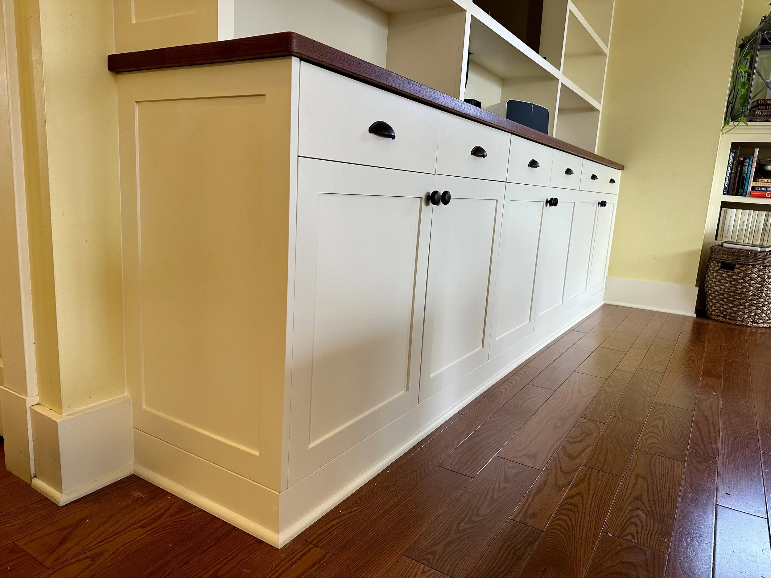 Wooden floor and a white kitchen cabinet with a dark brown countertop, with open shelving above it and a bookshelf on the right side.