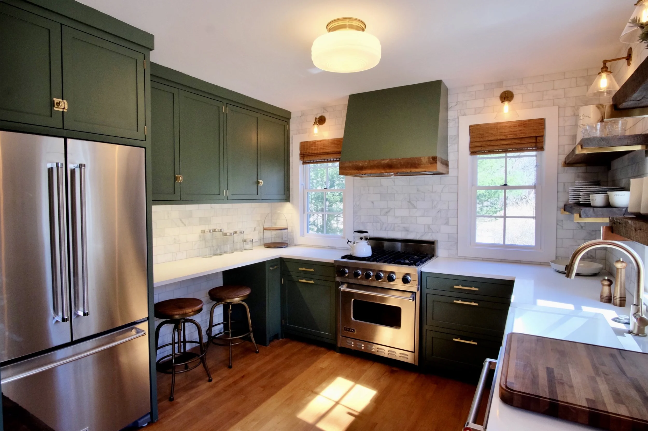 Modern kitchen with green cabinets, stainless steel refrigerator, white countertops, brick backsplash, wooden floors, and a stove with a range hood. Two small windows with wood blinds, open shelves with dishes, and a sink with a faucet.