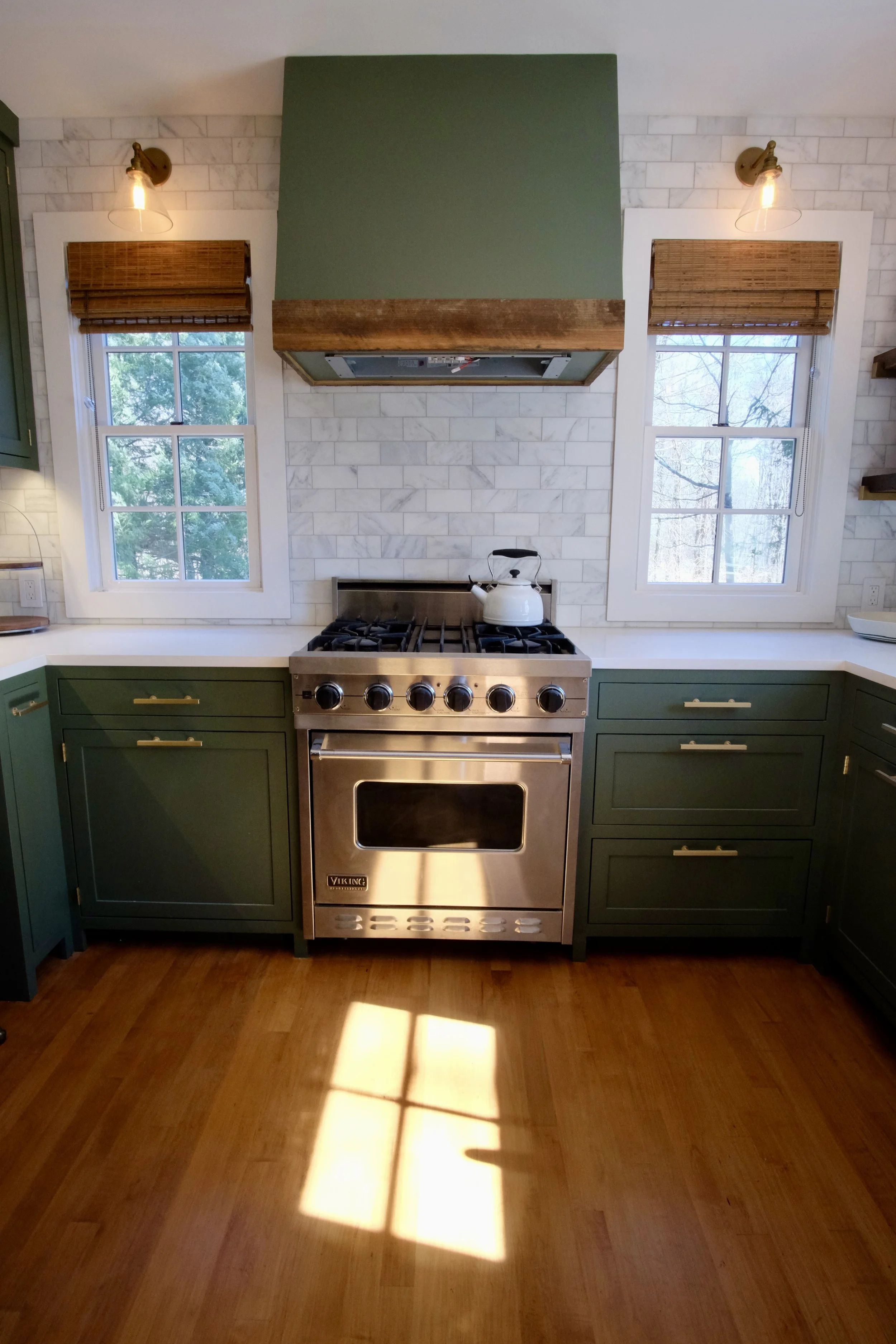 Kitchen with a stainless steel Viking stove, green cabinets, white countertops, two windows with bamboo shades, and a white teapot on the stove. Sunlight creates a window-shaped shadow on the wooden floor.