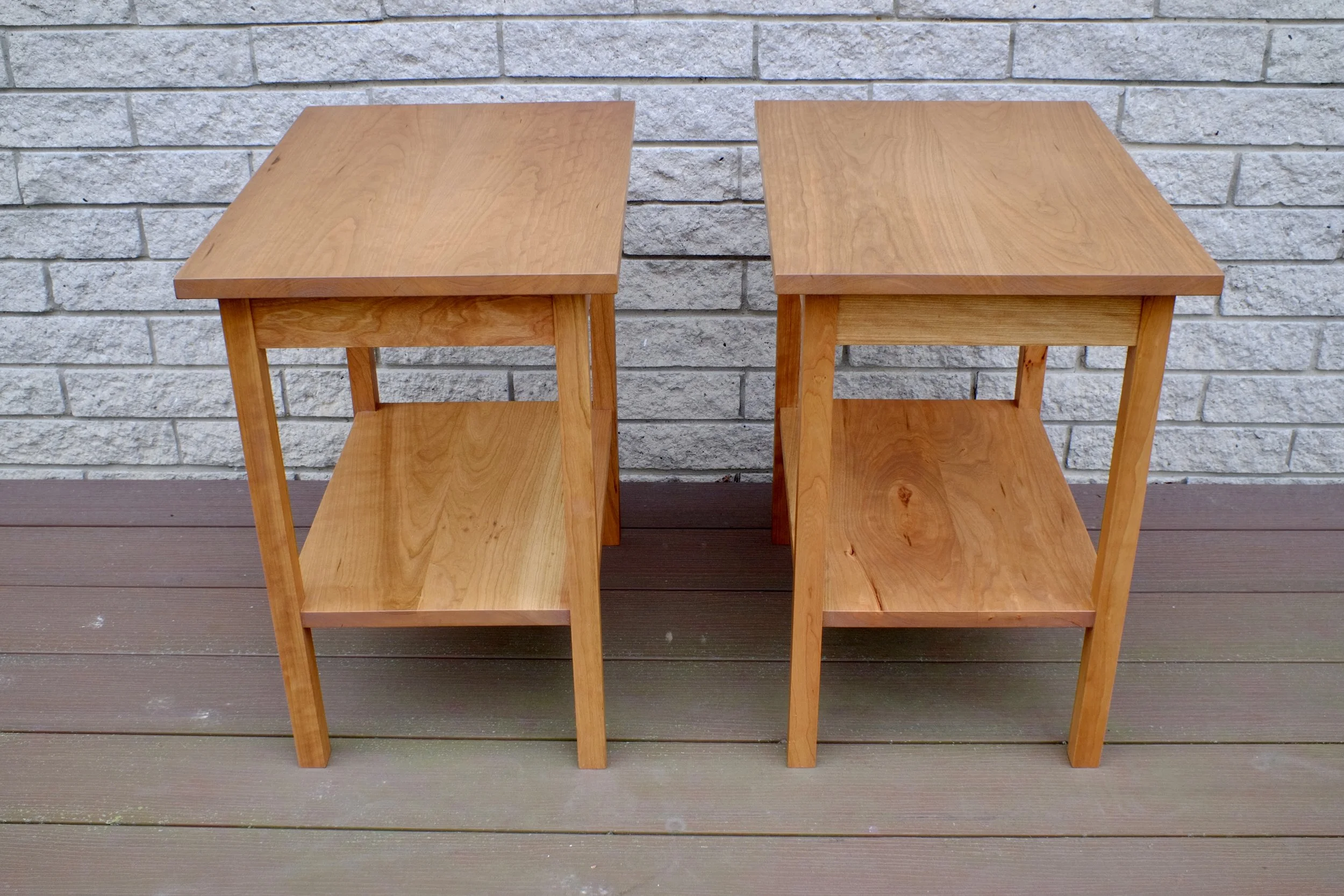 Two wooden side tables with shelves, placed outdoors on a wooden deck against a gray brick wall.