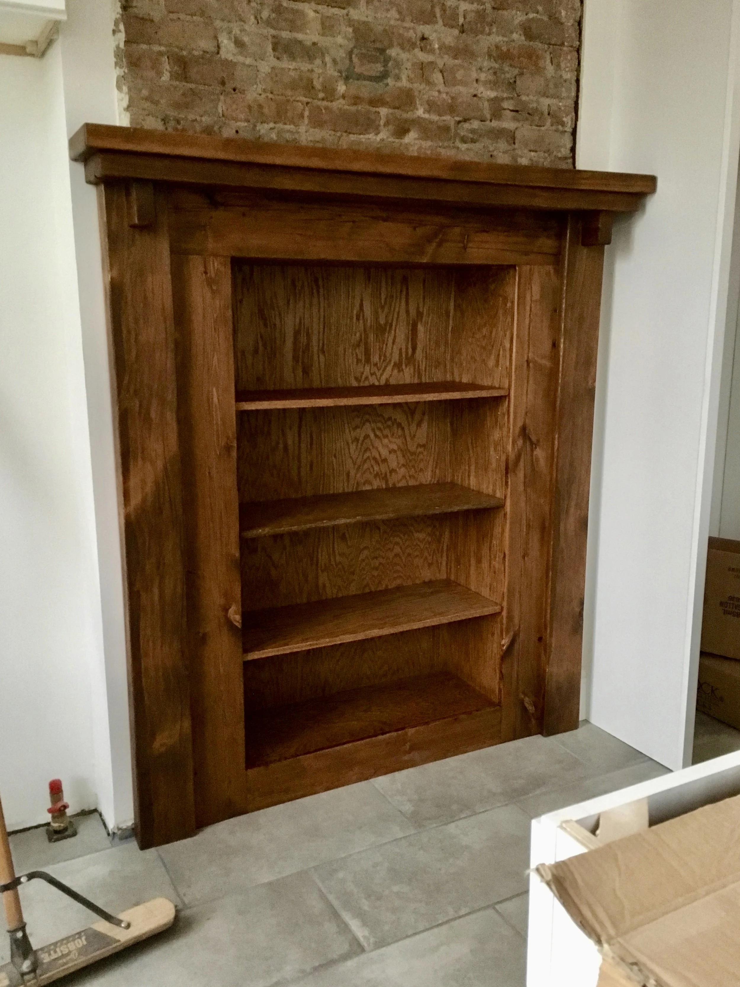 Empty wooden bookshelf with four shelves, placed against an exposed brick wall in a room with tiled flooring.