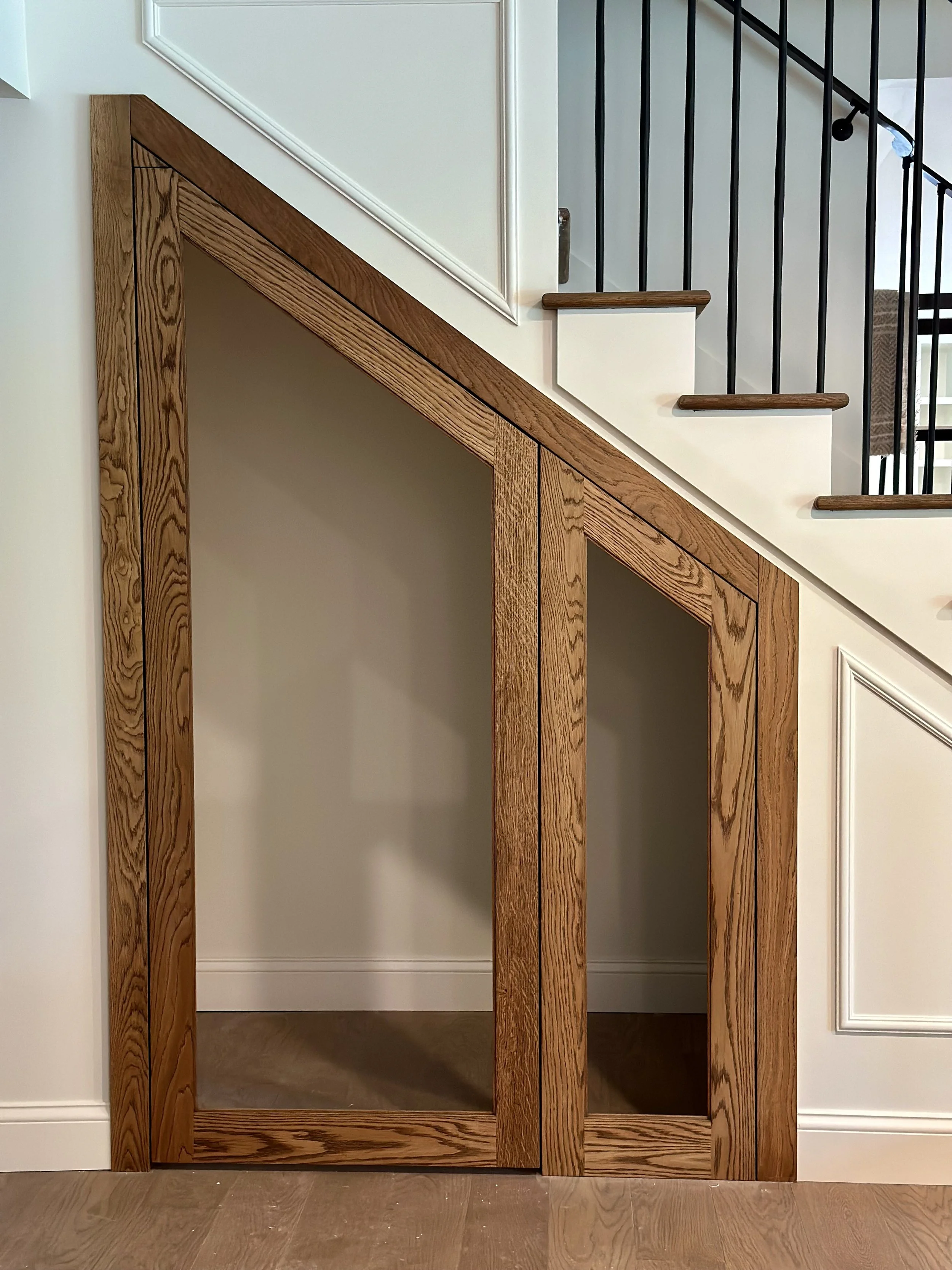 Custom interior wooden door with a glass panel, located underneath a staircase with black metal railing and wooden handrails, inside a modern home.