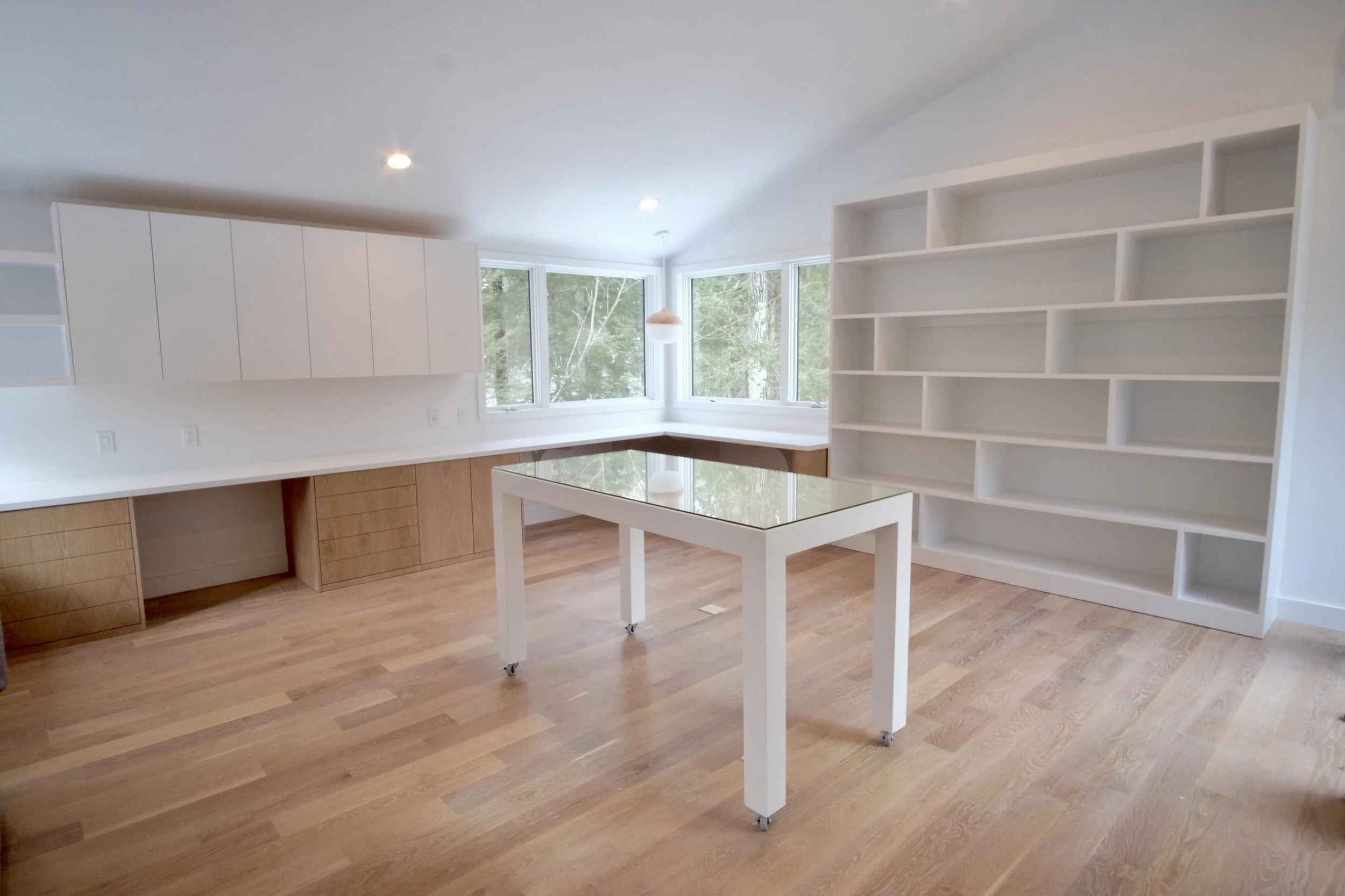 Empty room with wooden flooring, white walls, built-in white cabinets, a large window, a white bookshelf, and a white table with a glass top.