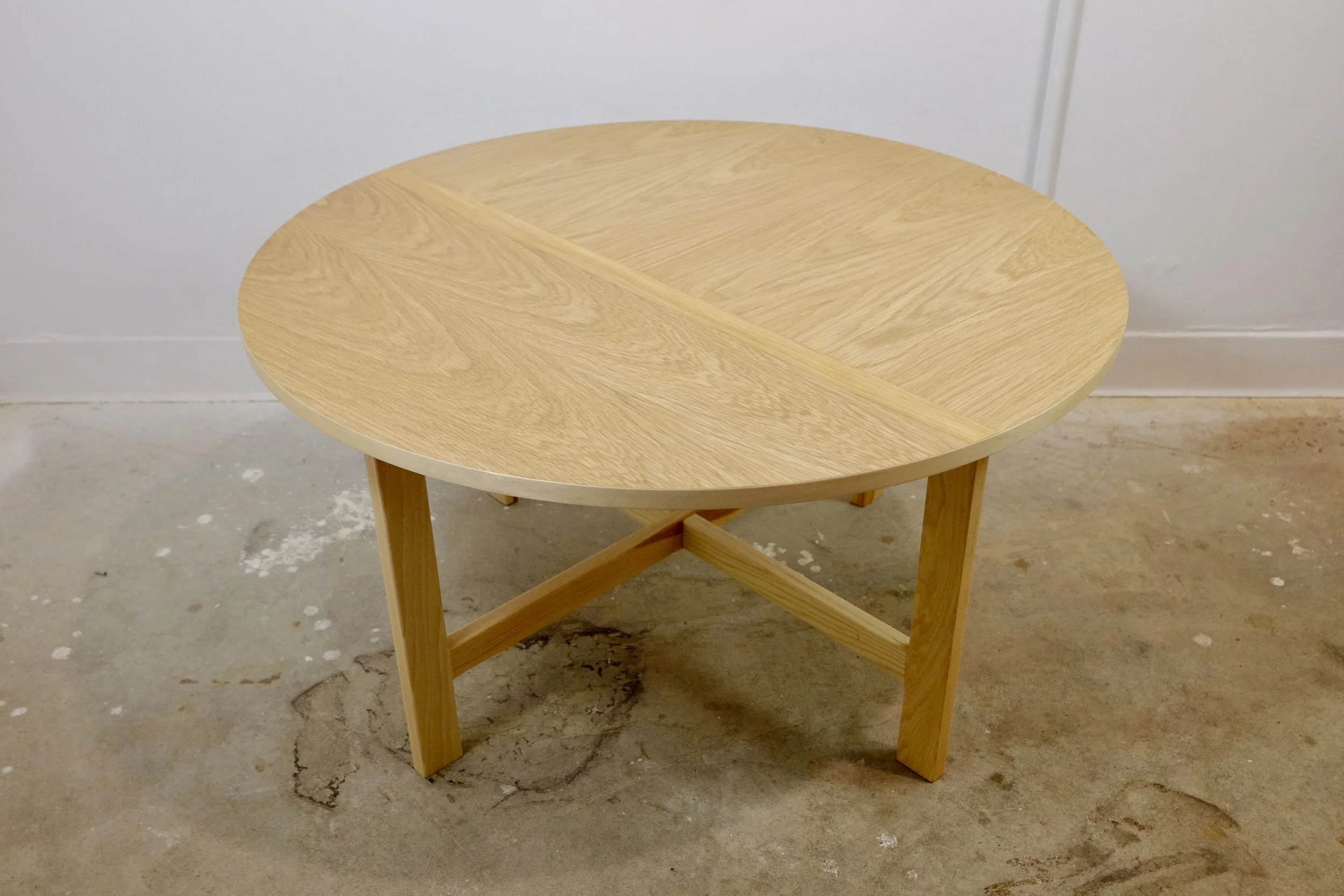 Round wooden table with a segmented top, supported by four legs and cross braces underneath, set against a plain white wall and a concrete floor.