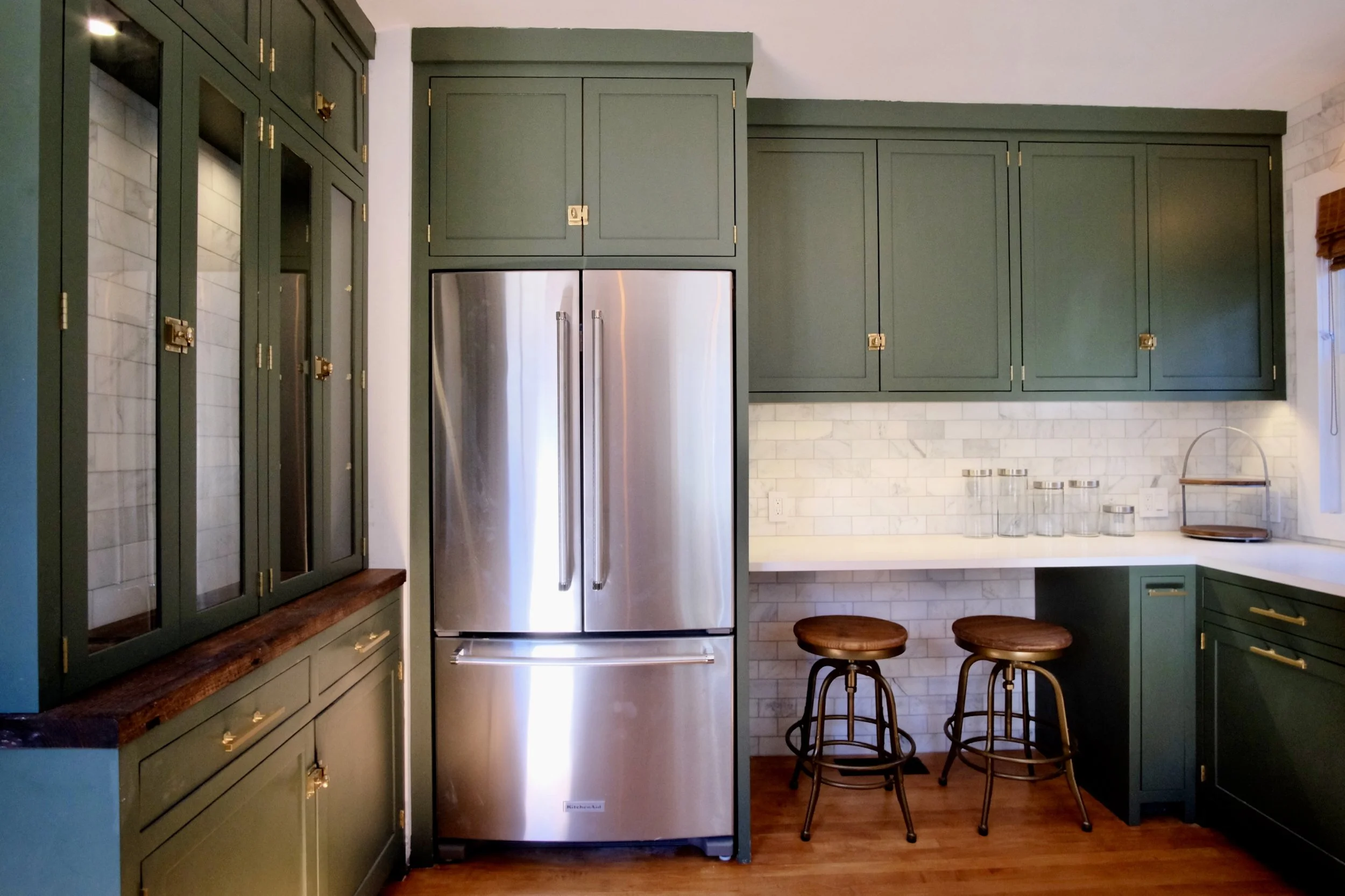 Kitchen with dark green cabinets, stainless steel refrigerator, white brick backsplash, wooden bar stools, and a white countertop.