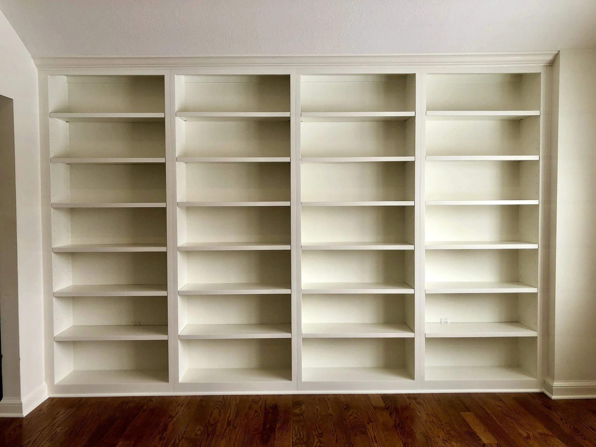 Empty white bookshelf with five columns of shelves against a beige wall and a wooden floor.
