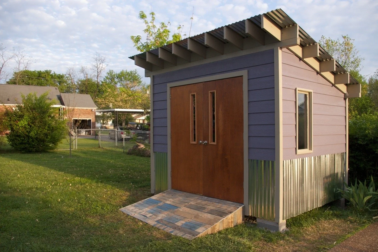 A small purple shed with light-colored trim, double wooden doors with narrow windows, a small window on the side, and a makeshift ramp at the entrance, situated in a grassy backyard with trees and neighboring houses in the background.