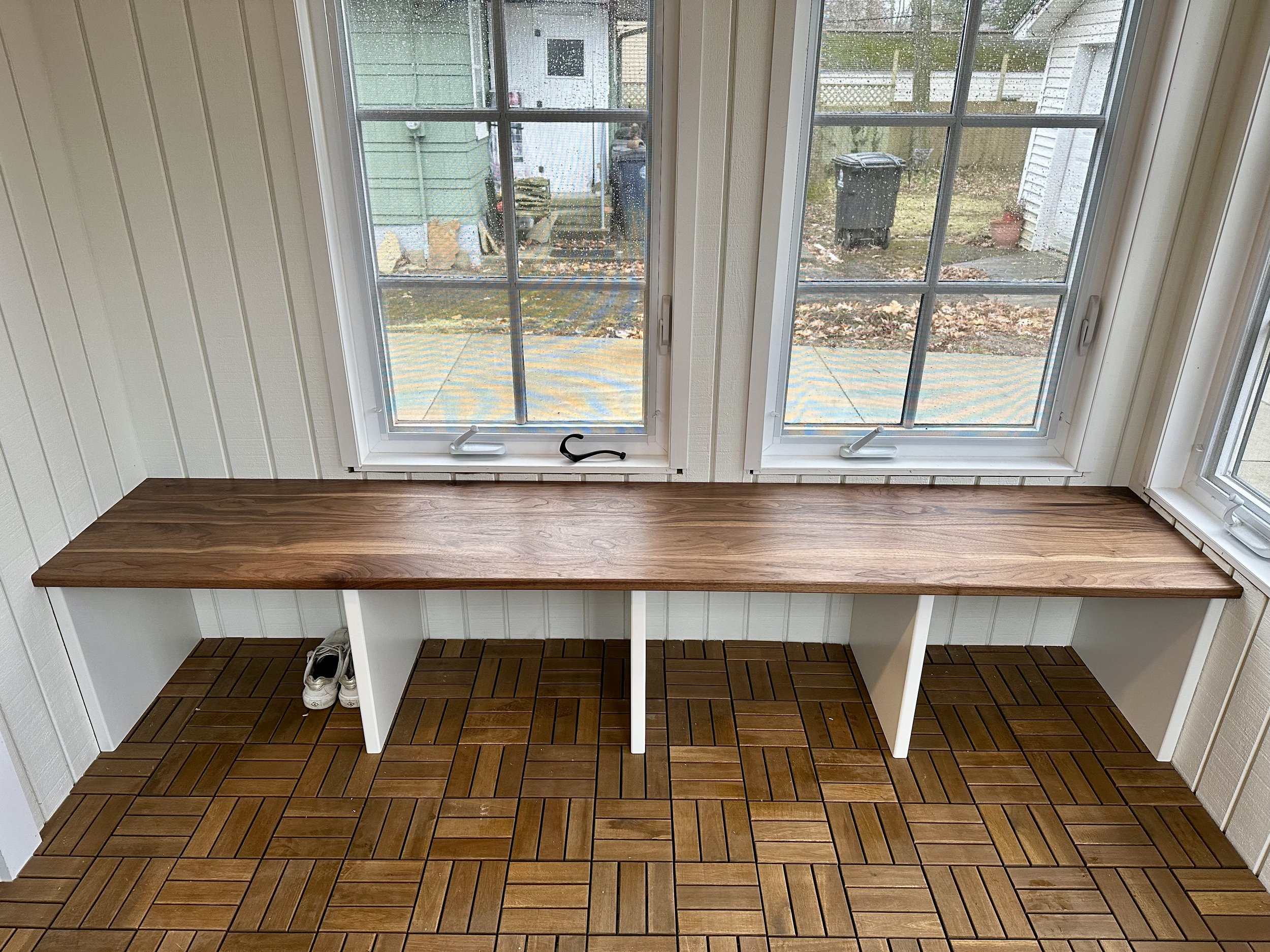 A wooden bench with white support legs situated in front of two large window panes in a room with white paneled walls and a wooden floor with a parquet pattern. A pair of white shoes is placed underneath the bench on the left side.