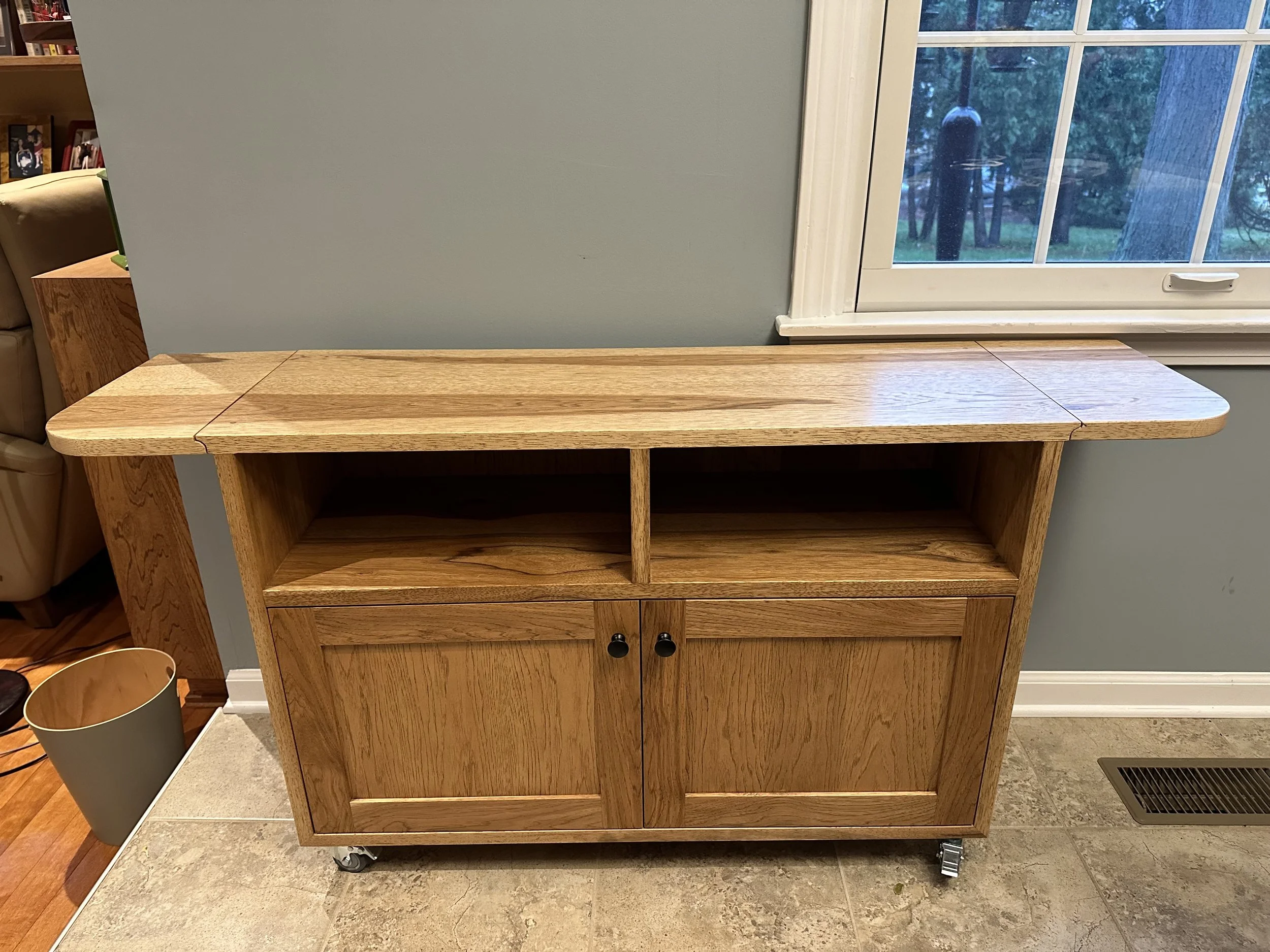 Wooden cabinet on wheels in front of a window with a blue wall behind it.