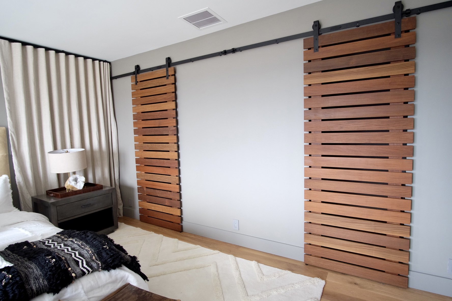 Interior of a bedroom with a wall decorated with two wooden panels on sliding tracks, a bedside table with a lamp and decorative objects, and a bed with a black and white blanket.