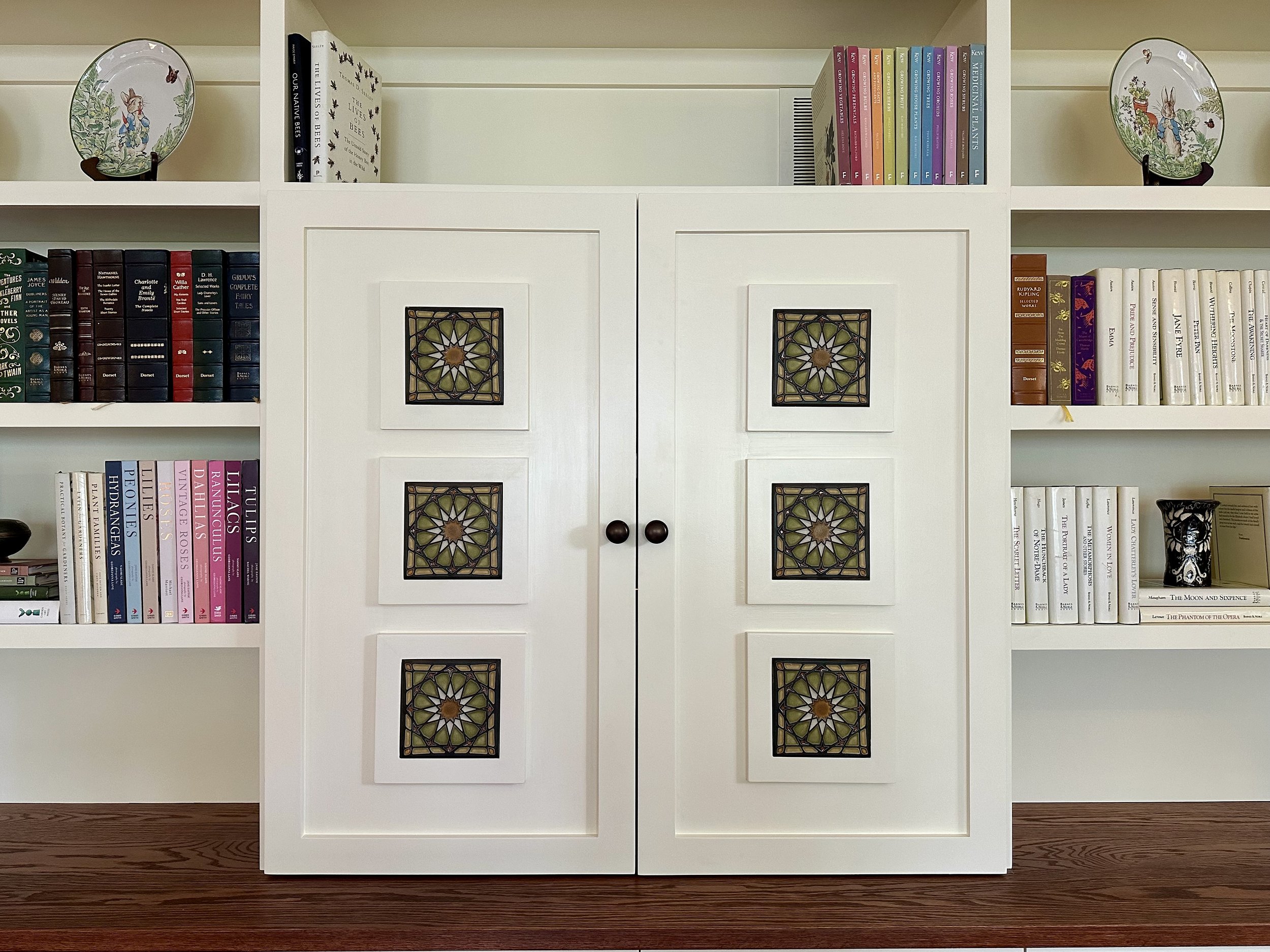 White bookshelf with decorative stained glass panels on cabinet doors, surrounded by colorful books and decorative plates.