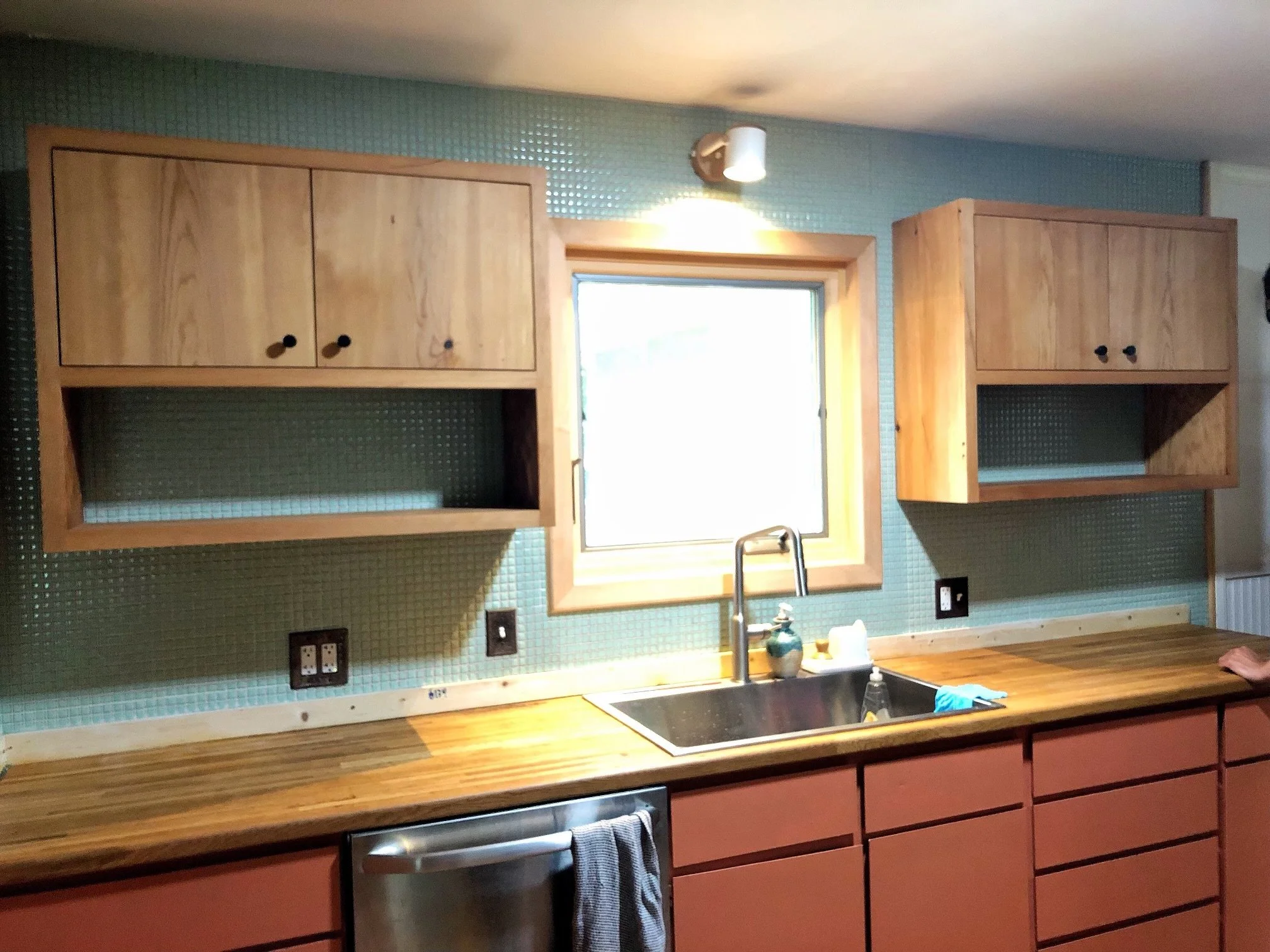 Wooden kitchen cabinets above a stone backsplash with a window in the center, a stainless steel sink, and a wooden countertop.