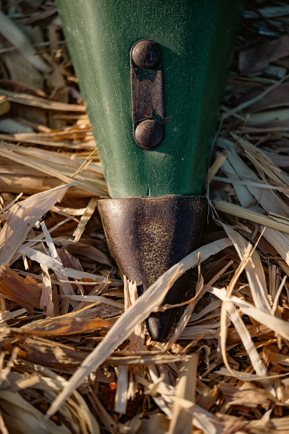 Close-up of a green metal fence post with rusted bolt, surrounded by dry straw or grass.