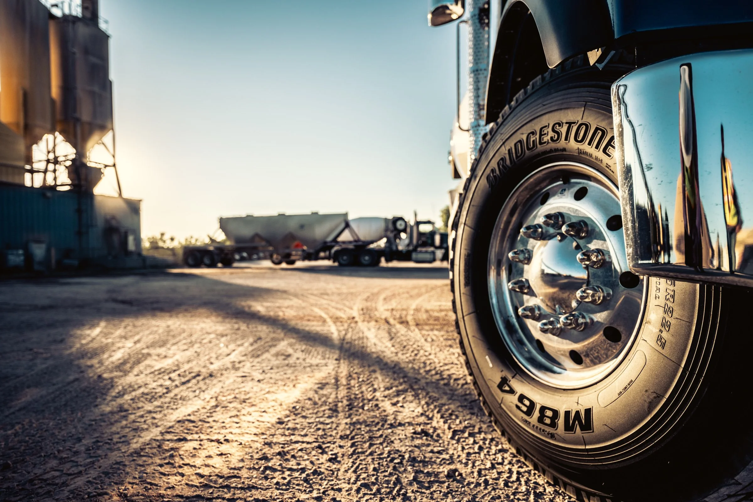 Close-up of a large truck wheel with Bridgestone tire on an industrial yard with several other trucks and trailers in the background during sunset.