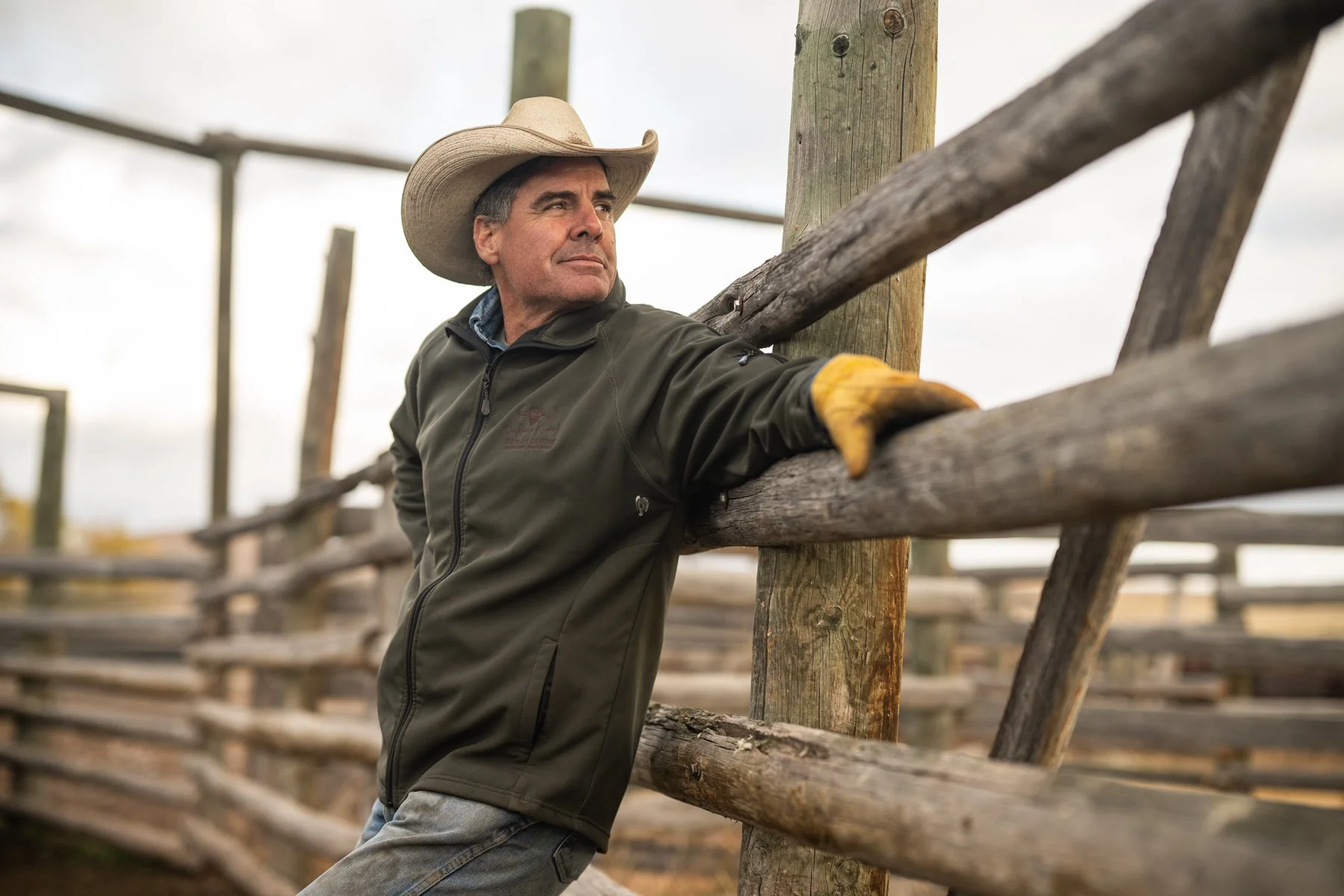 A man in a cowboy hat and jacket leaning on a wooden fence.