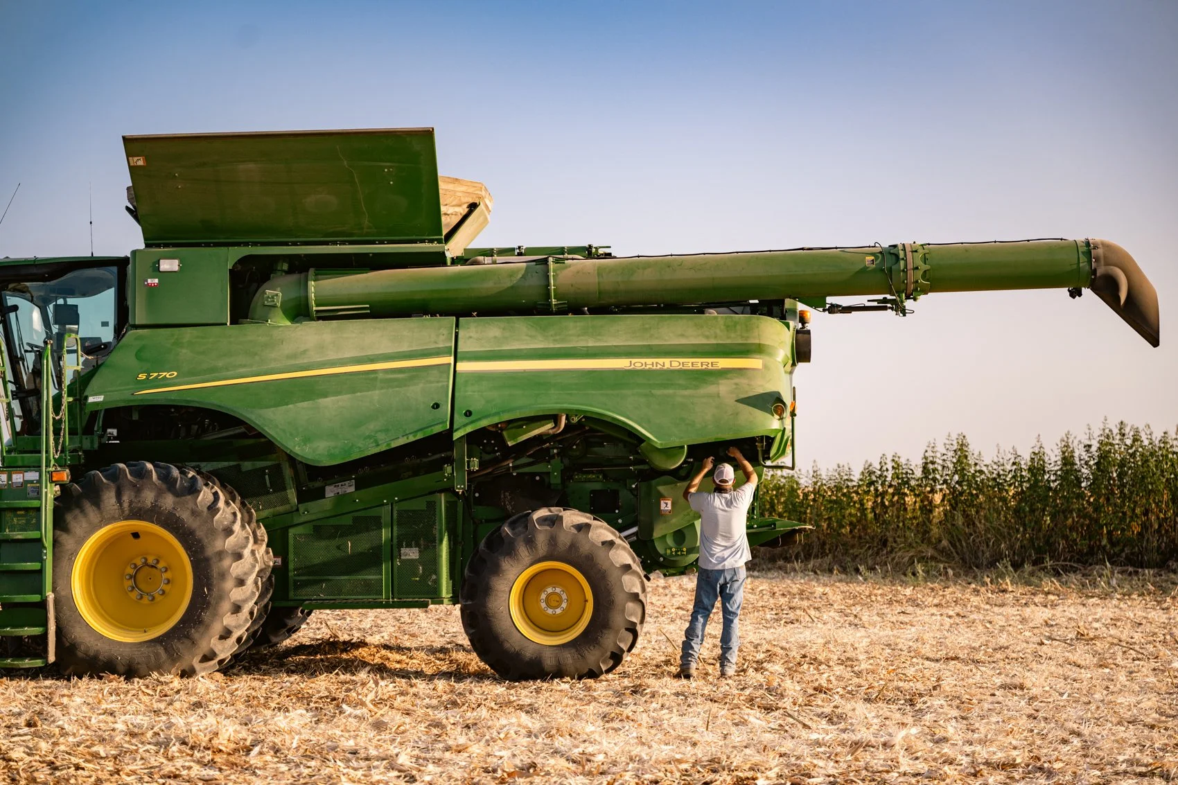 A person inspecting a large green John Deere combine harvester in a harvested field with a blue sky.