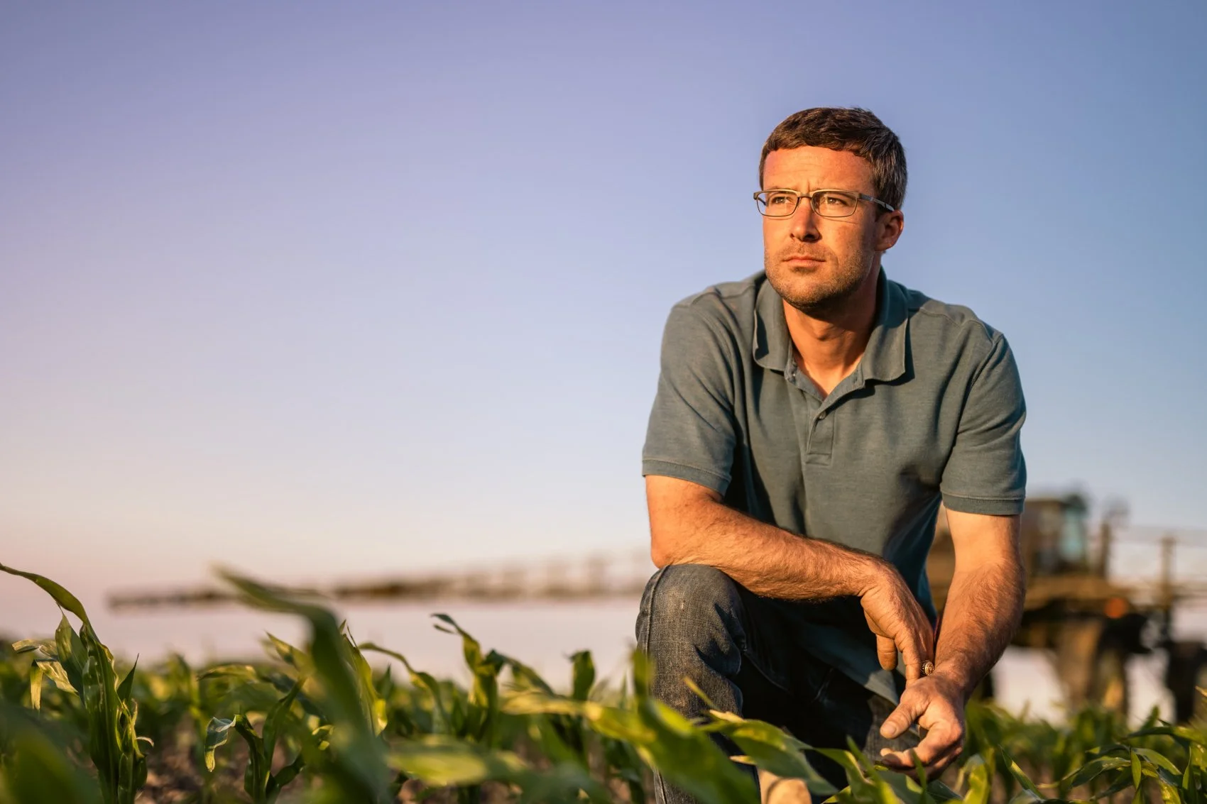 A man in glasses and a gray polo shirt crouches in a field of green crops, with a farm tractor blurry in the background, during late afternoon or early evening.