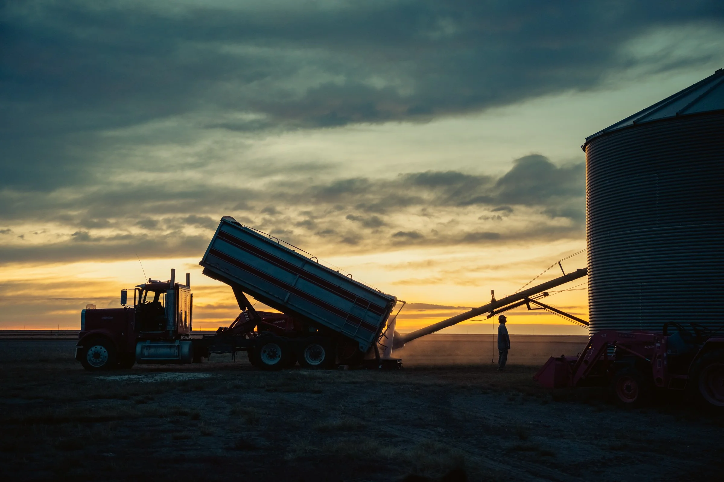 A farm scene at sunset showing a tractor with a raised trailer, a silo, and a person standing nearby, with a cloudy sky in the background.
