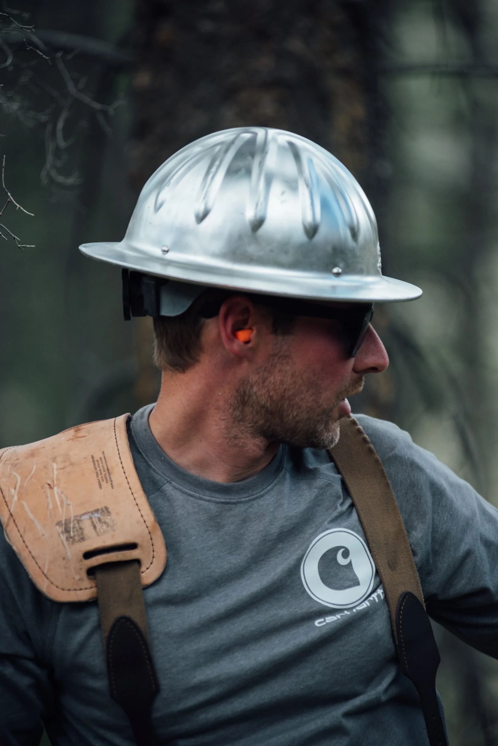 A man wearing a silver safety helmet, sunglasses, and ear protection, with a Carhartt T-shirt, outdoors in a wooded area.