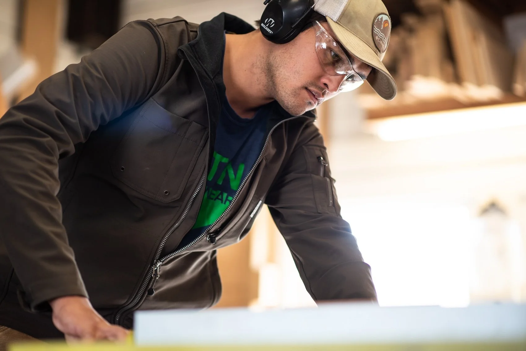 A man wearing safety glasses, a beige cap, and a brown jacket, working or studying at a table in a workshop or woodworking space.