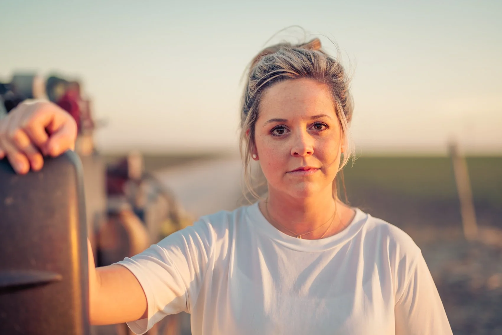 A young woman with blonde hair tied back, wearing a white t-shirt, taking a selfie outdoors at sunset, with a blurred background of green fields and a bicycle.