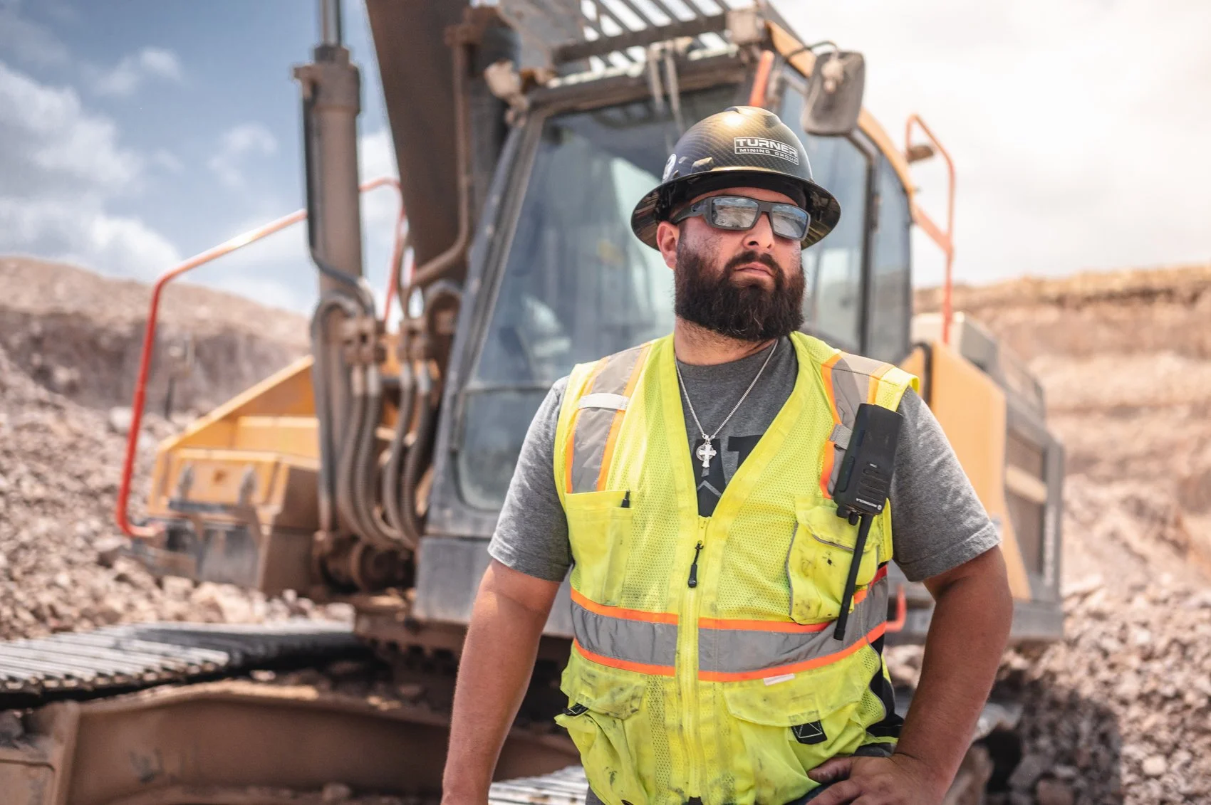 A construction worker with a beard wearing safety glasses, a helmet, a yellow safety vest, and a dark t-shirt stands in front of a large excavator at a mining site with rocky terrain under a partly cloudy sky.