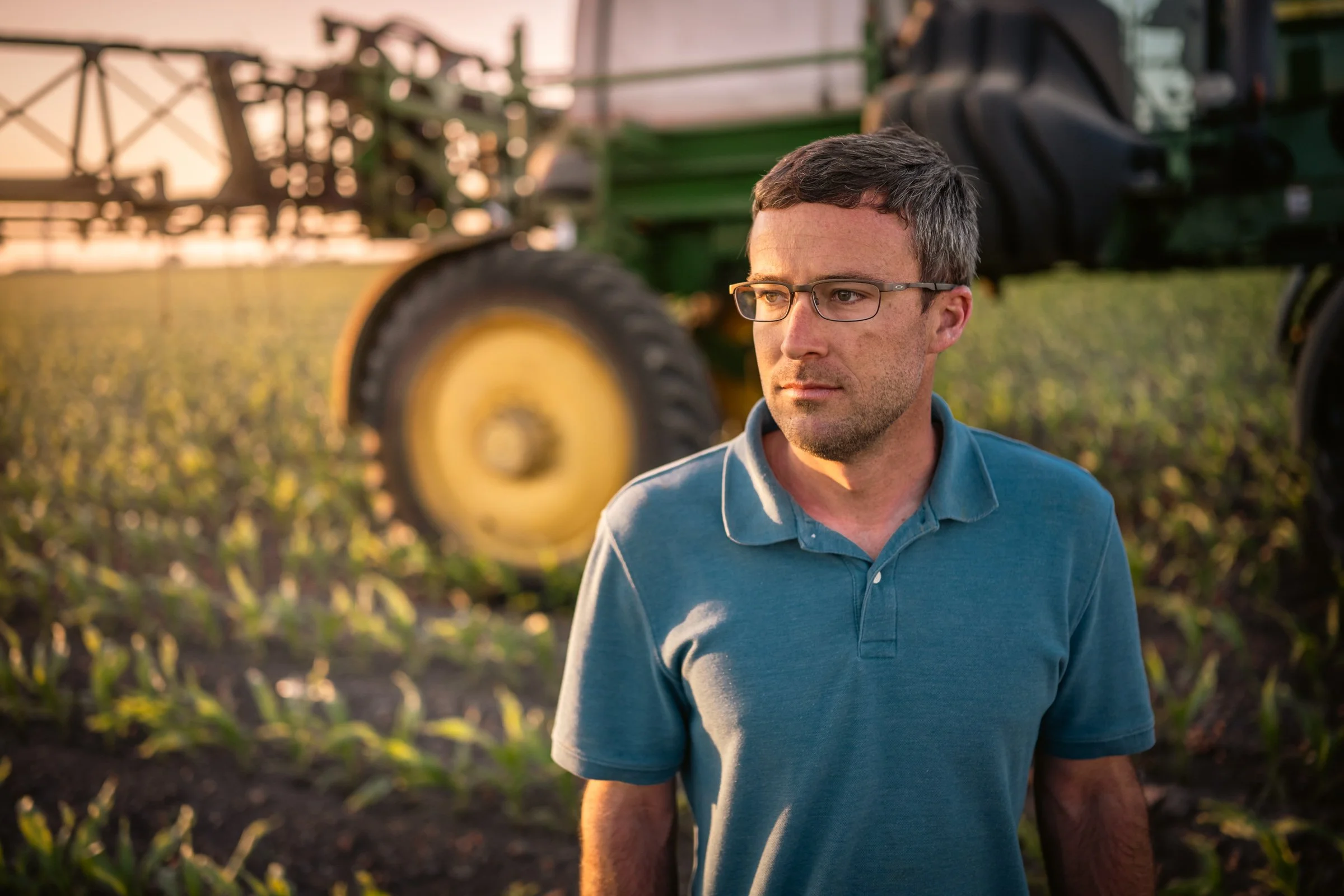 A man wearing glasses and a blue polo shirt stands in a field with a tractor in the background during sunset, looking to the side.