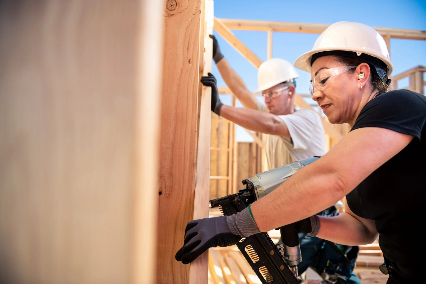 Two women wearing white safety helmets, safety glasses, and gloves using power tools to work on wooden framing at a construction site. One woman is in the foreground, operating a nail gun, while the other is in the background, securing a wooden beam.