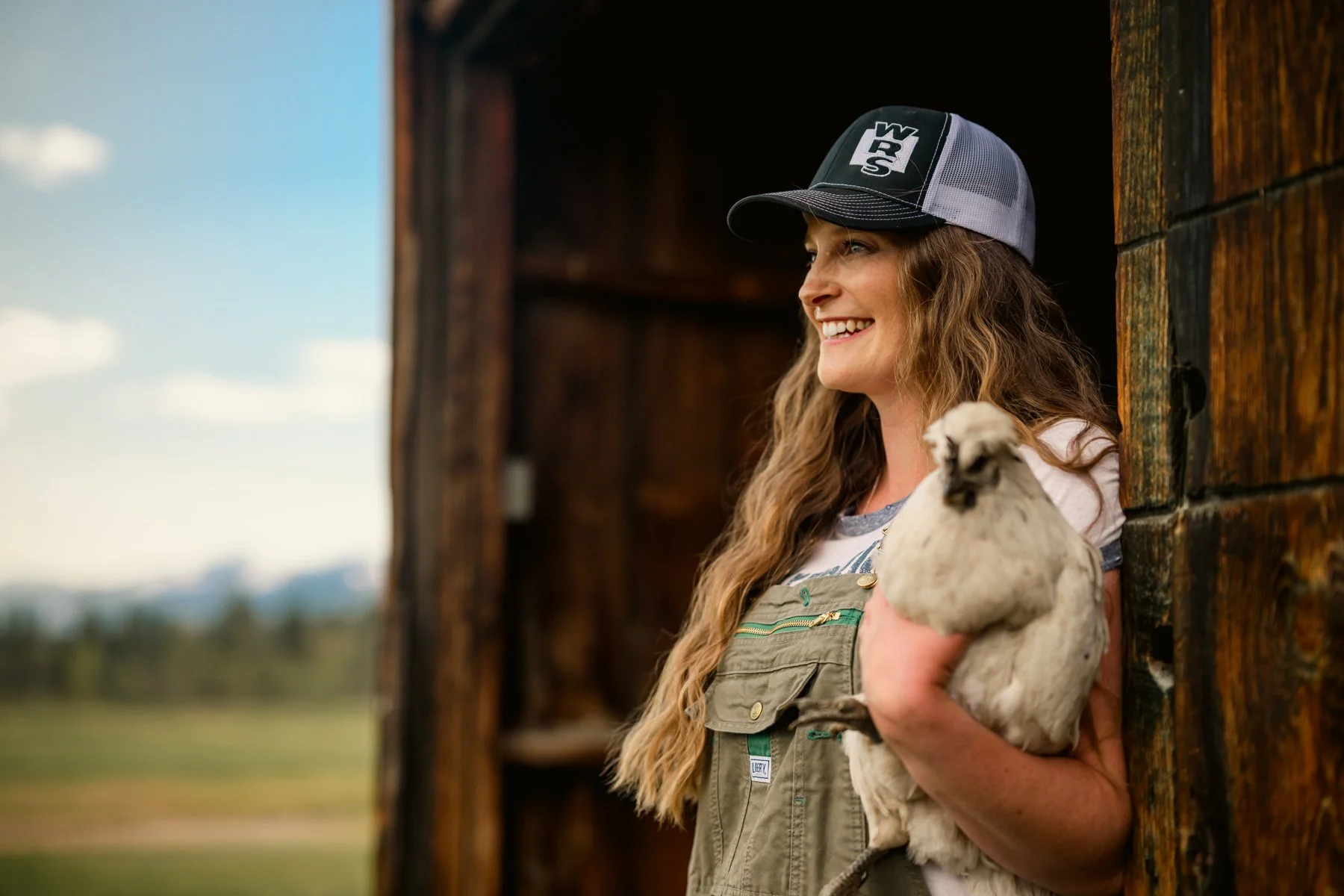 A woman with long, curly hair smiling and wearing a trucker hat and overalls, holding a small llama, standing outside a rustic wooden barn with a rural landscape behind her.
