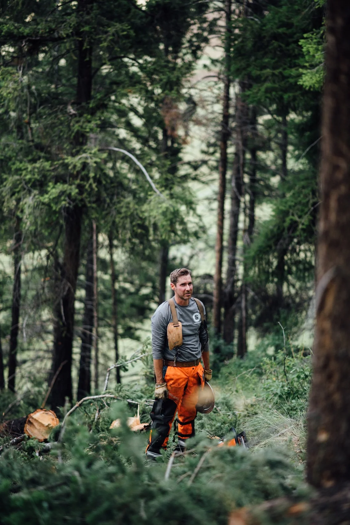 A man in outdoor gear stands in a forest with trees and greenery around him, holding a helmet and wearing gloves.
