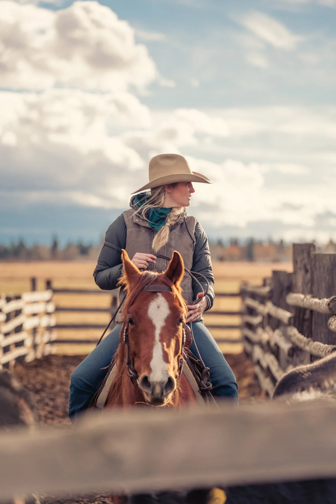 A woman wearing a cowboy hat and gray jacket riding a horse on a farm with fences and open fields under a partly cloudy sky.