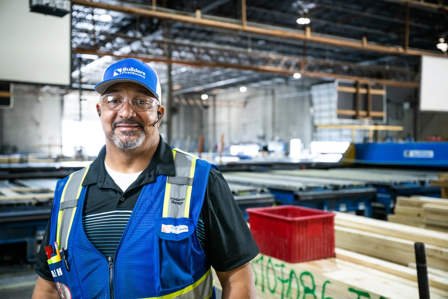 A man wearing a blue Builder FirstSource cap, safety glasses, a headset, and a blue safety vest at a construction supply warehouse.