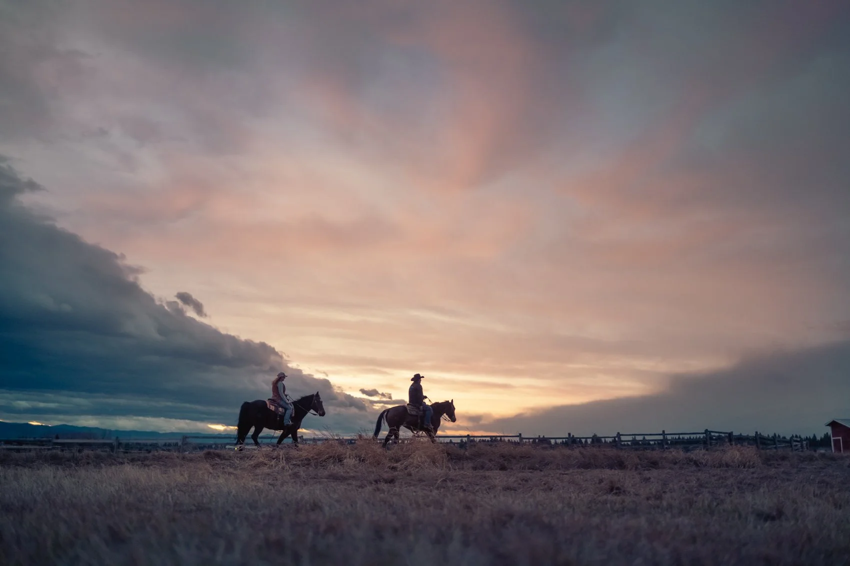 Two people riding horses across a grassy field at sunset, with cloudy sky and a fence in the background.