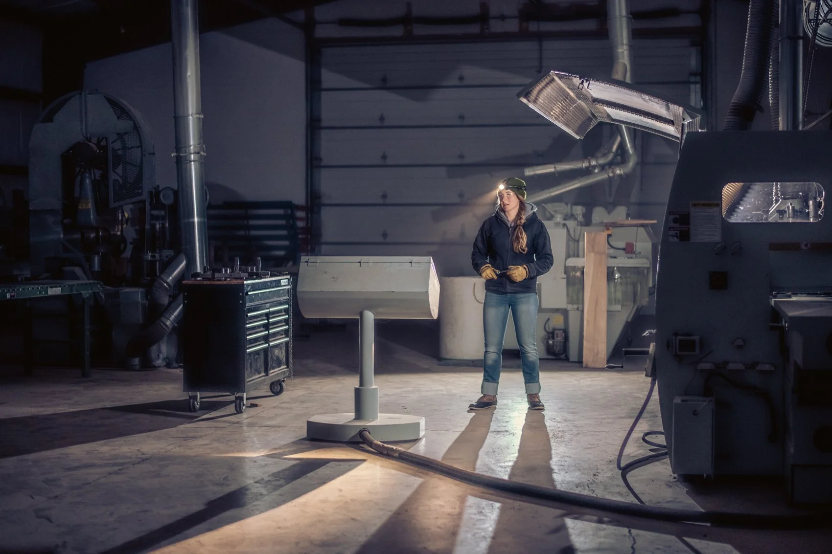 A woman in a dark jacket, jeans, and a headlamp stands in a workshop or industrial setting, holding a tool, surrounded by various machinery and equipment.