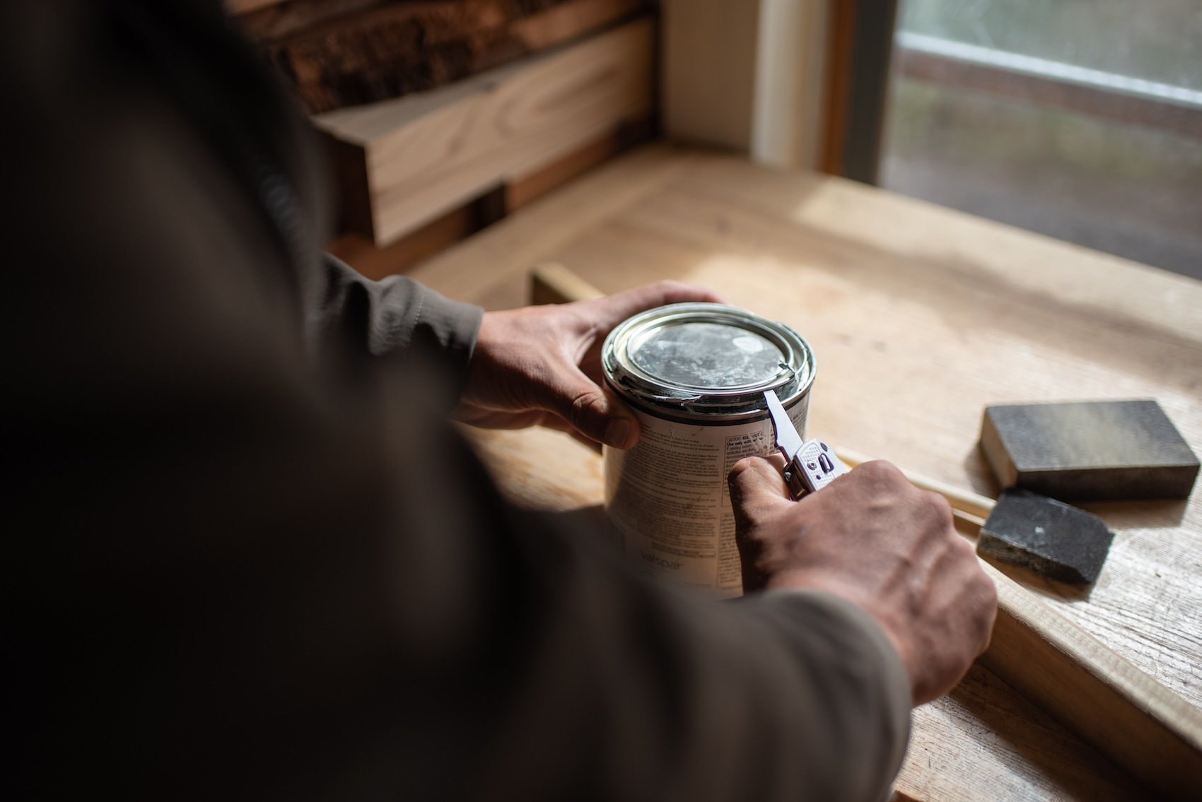 Person opening a can of paint with a can opener on a wooden workbench near a window.