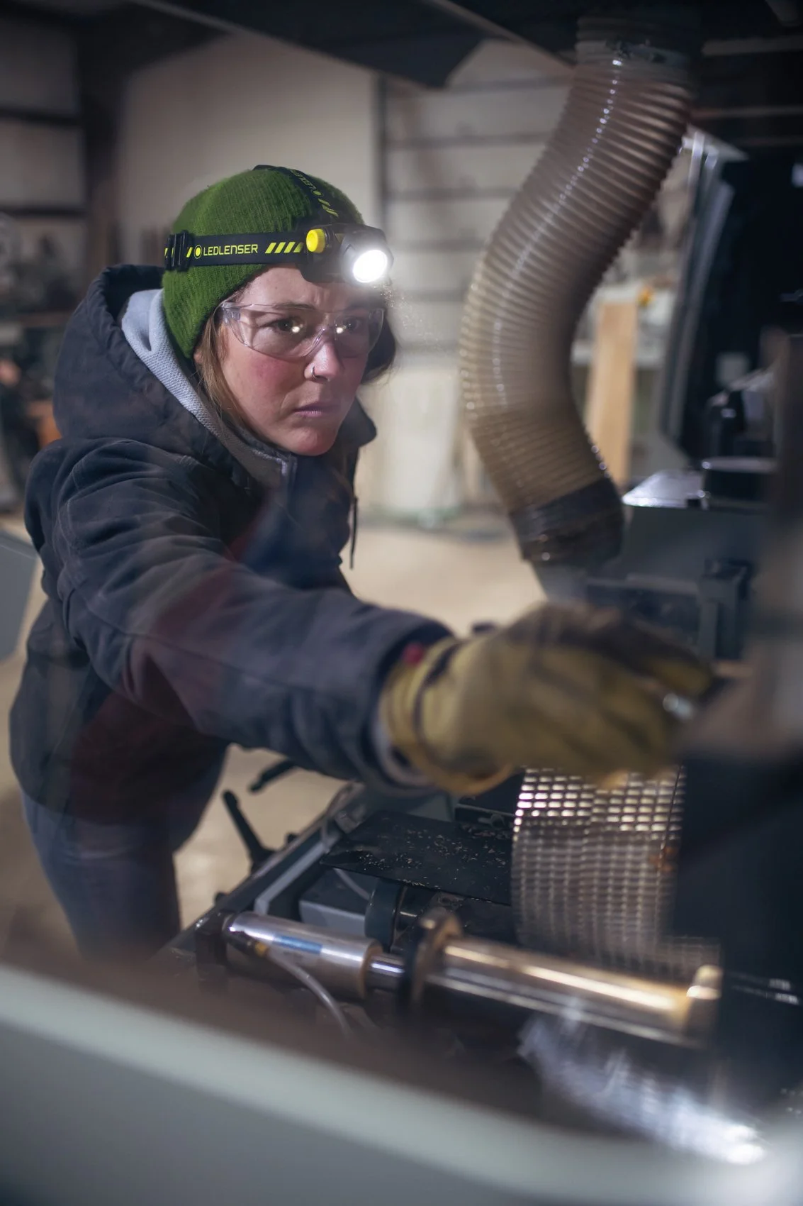A woman wearing safety goggles, a green beanie with a headlamp, a dark jacket, and work gloves is operating a machine in a workshop or garage.