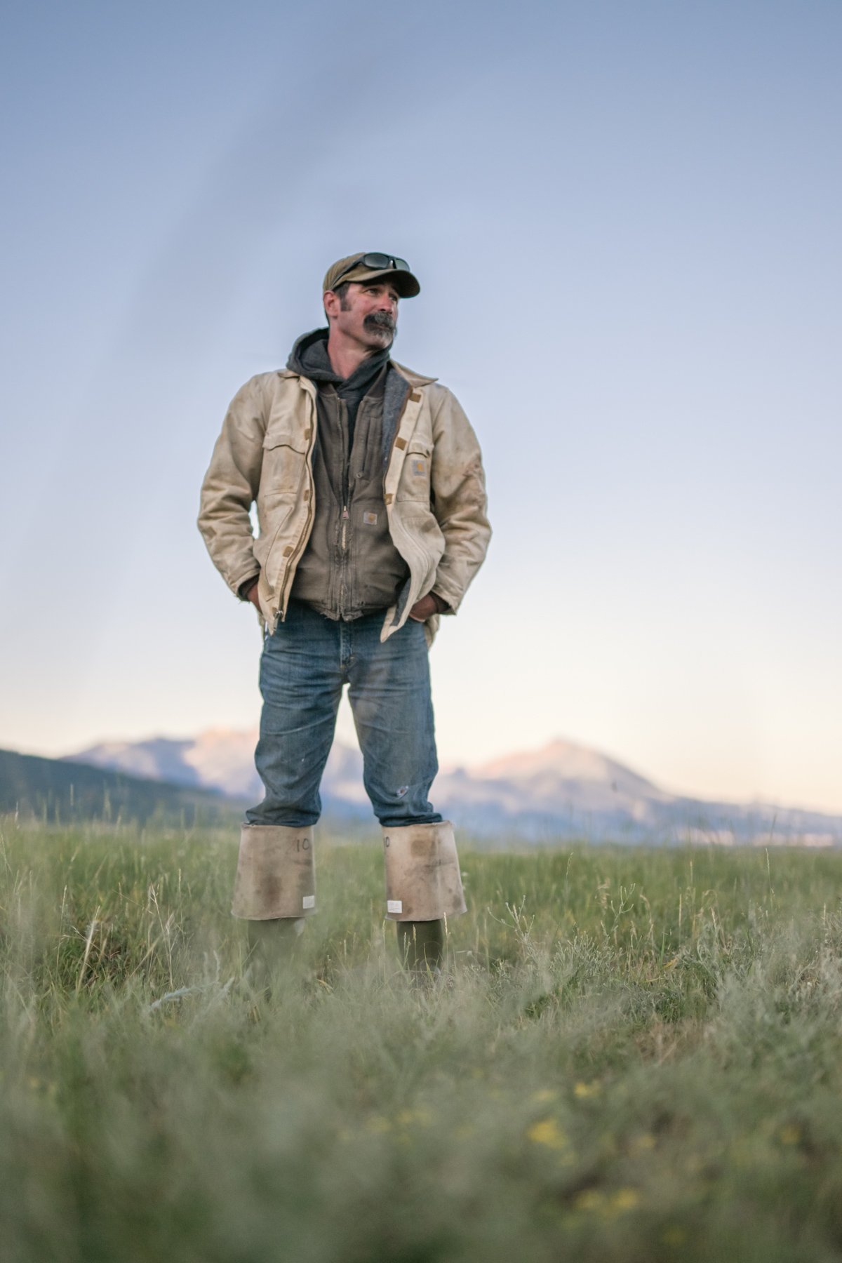 A man standing outdoors in a grassy field with mountains in the background during daytime, wearing a beige jacket, dark hoodie, jeans, and rubber boots.