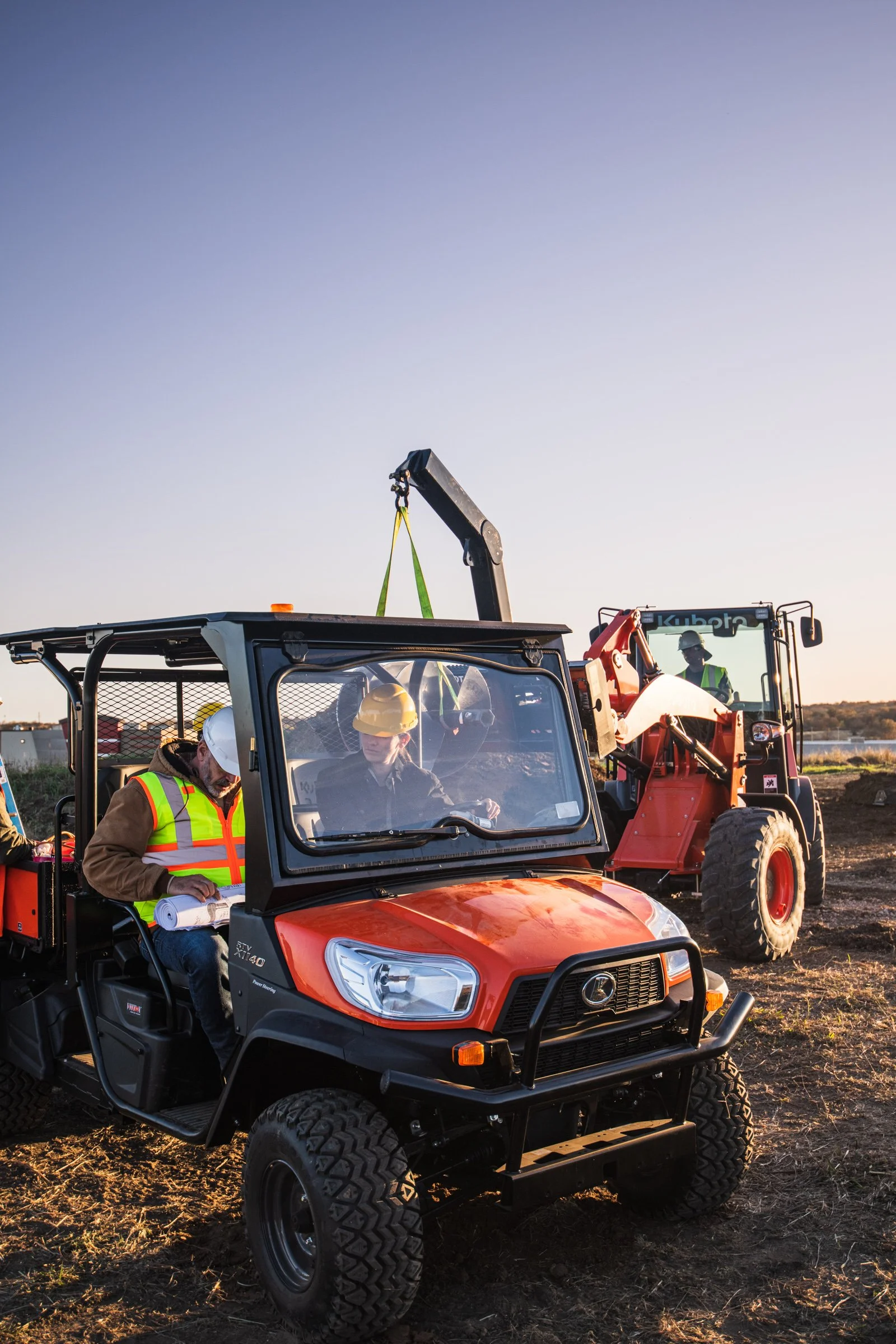 Construction workers wearing safety helmets and vests operate machinery on a construction site during daylight, including a utility vehicle and a loader.