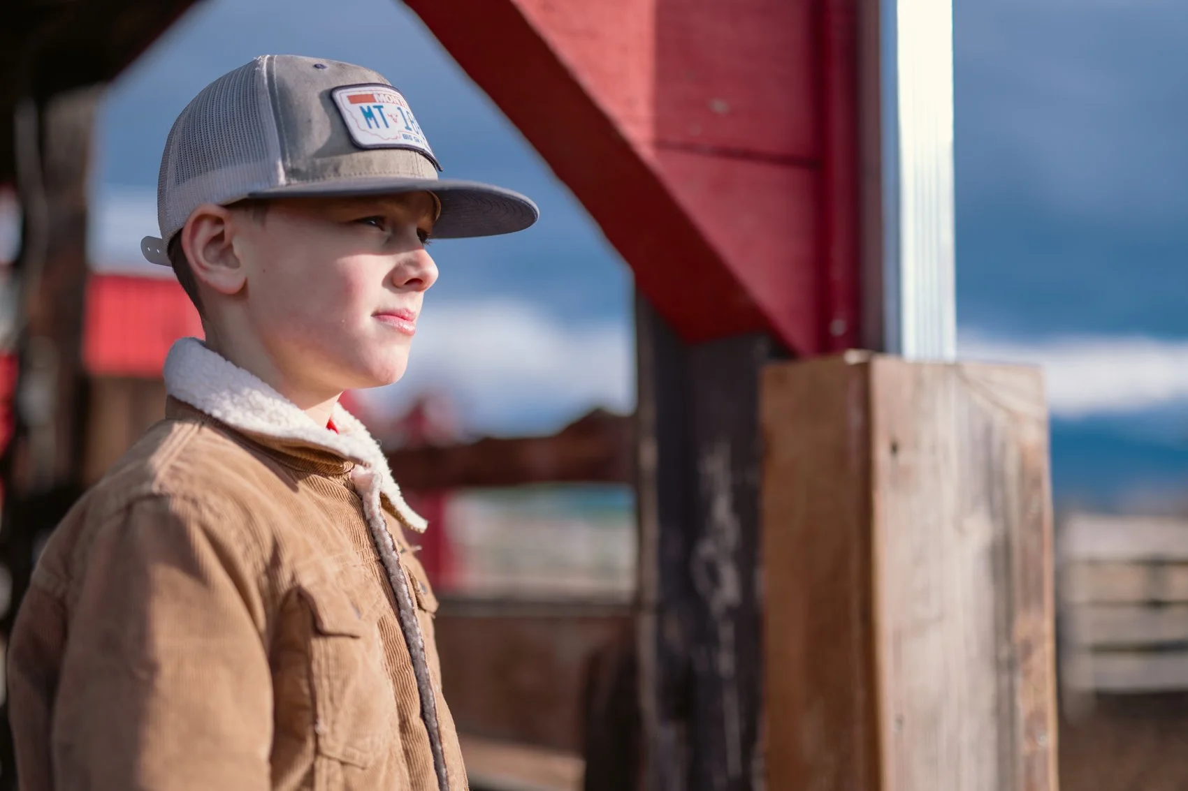 A young boy wearing a beige jacket and a baseball cap looks into the distance outdoors with a cloudy sky and wooden structures in the background.
