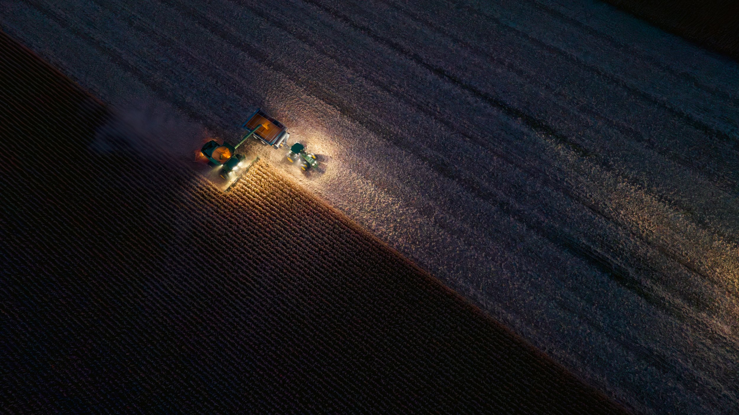 An aerial view at night of a tractor harvesting a field, with bright lights illuminating the machinery and the surrounding crops.