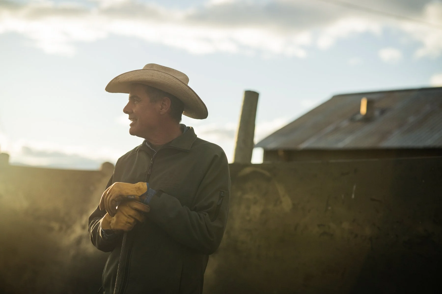 A man wearing a cowboy hat and brown gloves, standing outdoors near a barn at sunset.